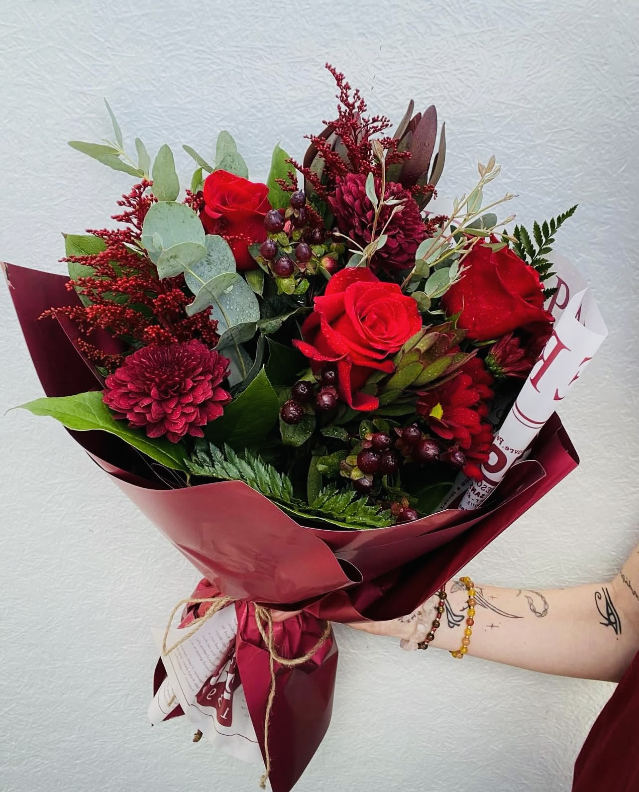 Hand-tied bouquet with red roses, red chrysanthemums, and berries wrapped in dark red paper.