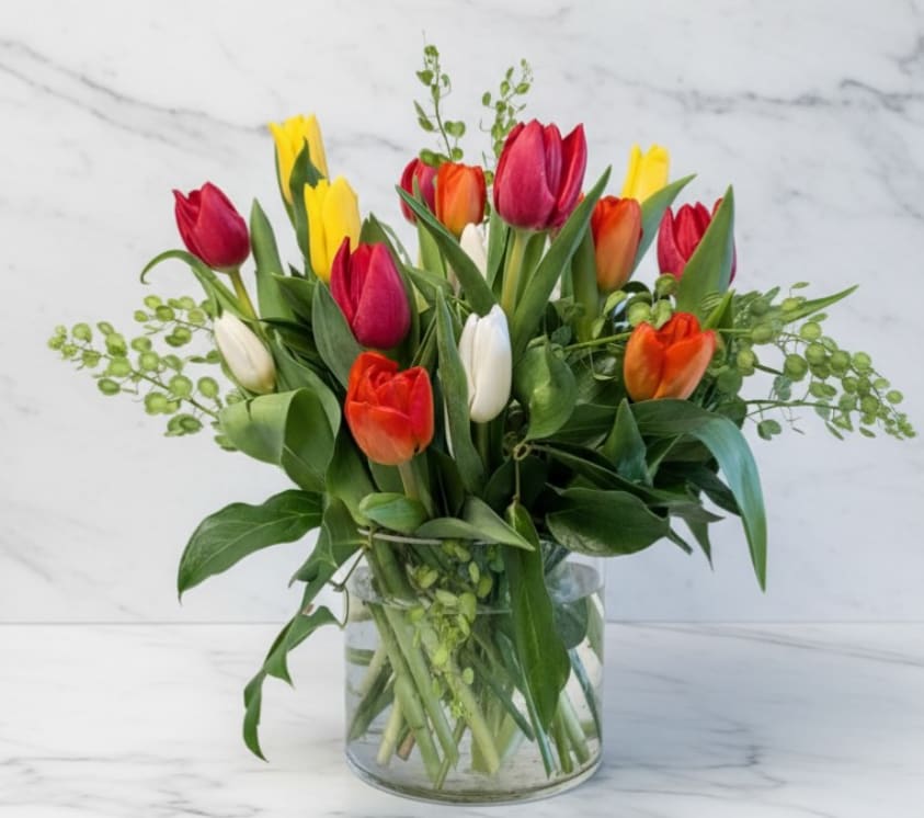 Spring Confetti Tulips - A cheerful mix of vibrant tulips in shades of red, yellow, orange, and white arranged in a clear glass vase with fresh greenery. This bright, seasonal arrangement captures the joy of spring and brings effortless color and elegance to any space. Perfect for birthdays, celebrations, or simply brightening someone’s day.