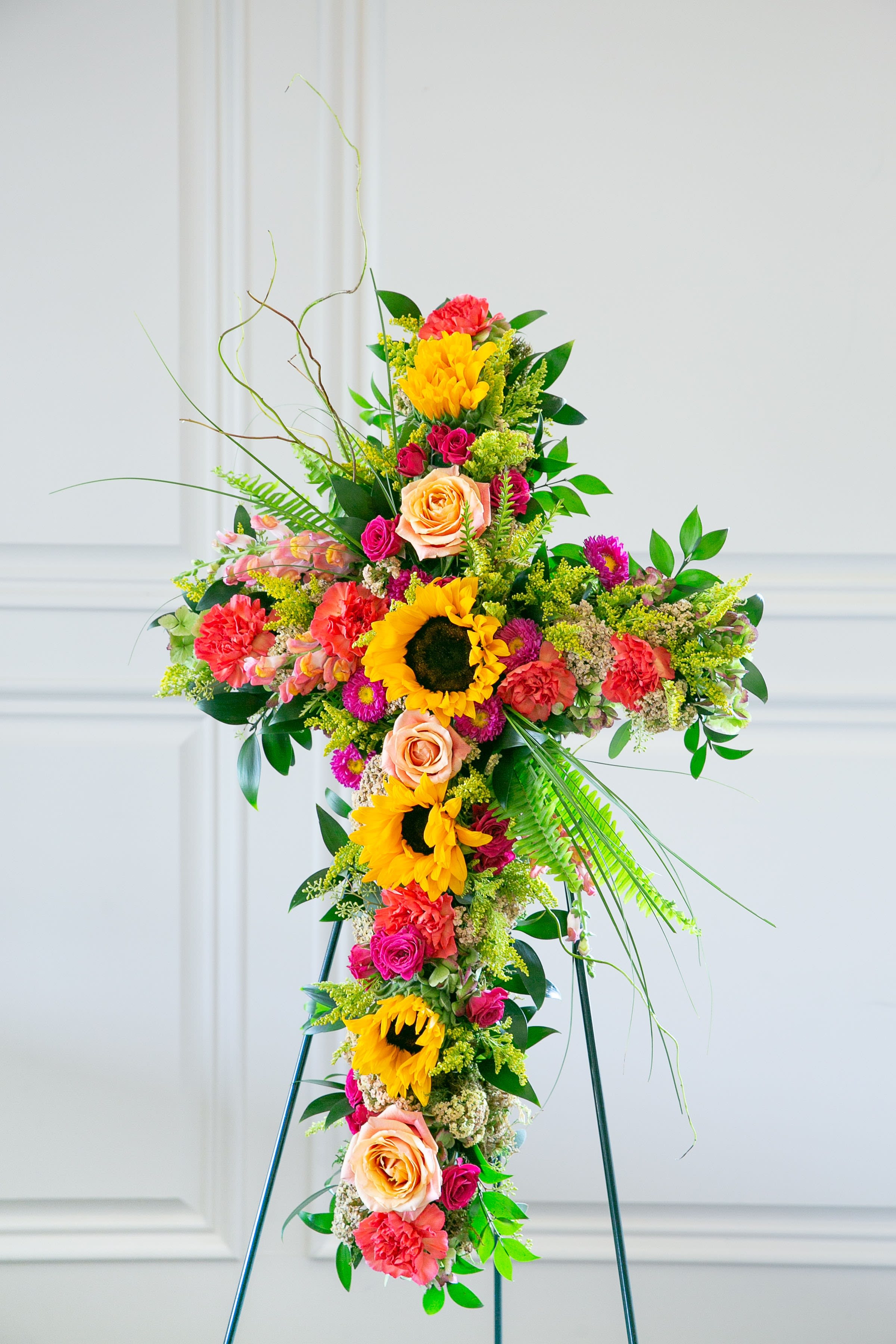 Wild Meadow Cross – Celebration of Life Tribute  - Bright, natural, and full of heartfelt warmth, the Wild Meadow Cross is a floral tribute designed to celebrate a life remembered with love and joy.  This standing cross features a vibrant blend of sunflowers, garden roses, seasonal wildflowers, and fresh greenery arranged with natural movement and organic texture. The warm tones of golden yellow, coral, peach, and soft pink create a garden-inspired design that feels uplifting and full of life.  Symbolizing faith, remembrance, and the beauty of cherished memories, this cross offers a meaningful tribute during funeral services, memorial gatherings, or celebration-of-life ceremonies.  Designed on a standing easel, the cross creates a graceful focal point while honoring the spirit and individuality of the loved one being remembered.  Each piece is handcrafted by the designers at ImpeKable Flowers in DeLand, using carefully selected seasonal blooms sourced from premium growers.  Flower varieties may vary depending on seasonal availability while maintaining the same wildflower-inspired color palette and design style.