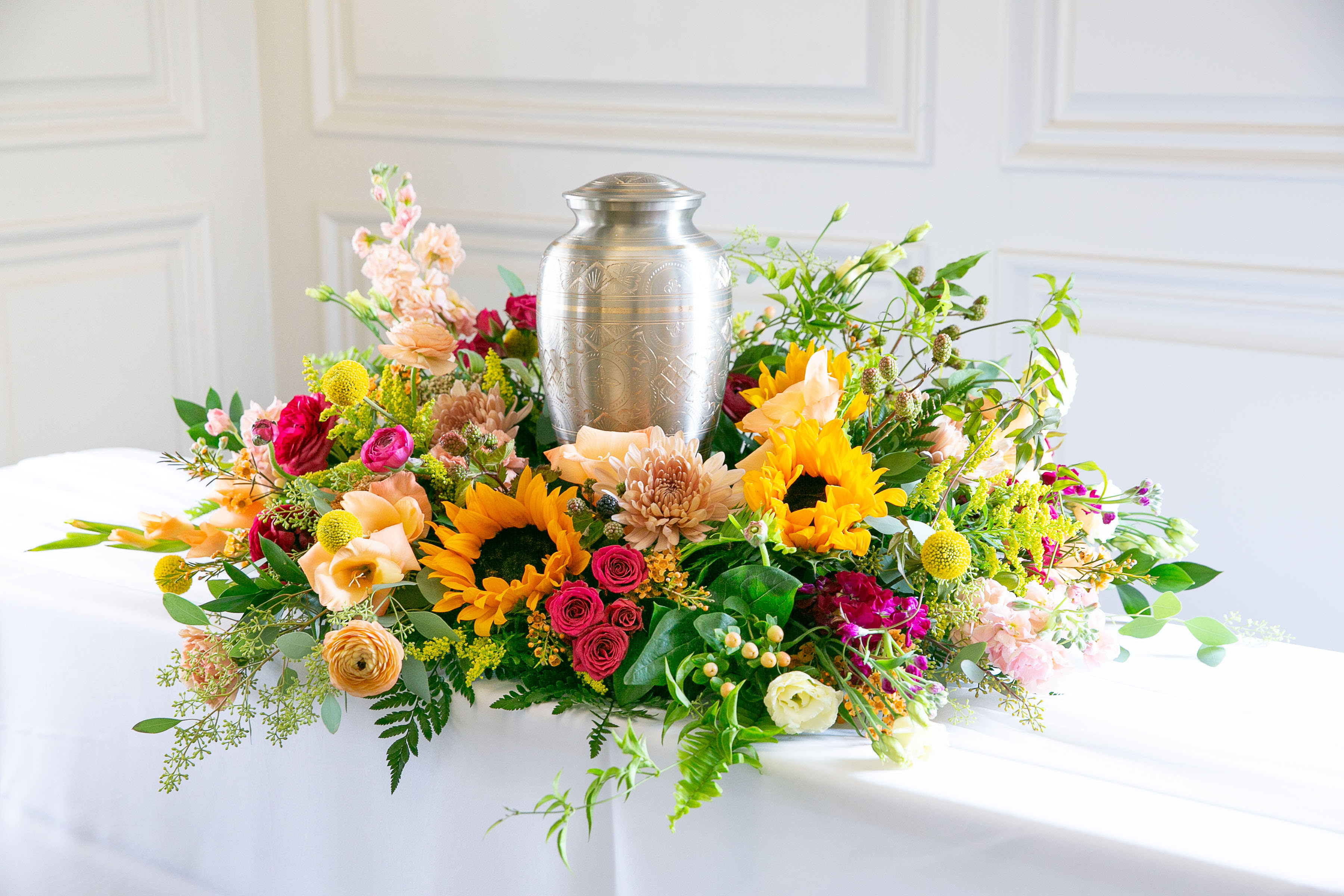 Wild Meadow – Cremation Urn Wreath - Warm, natural, and filled with heartfelt beauty, the Wild Meadow Cremation Urn Wreath is a floral tribute designed to honor and celebrate a life remembered with love.  This garden-inspired design features radiant sunflowers, garden roses, seasonal wildflowers, and fresh greenery arranged in a circular wreath surrounding the urn. The vibrant tones of golden yellow, coral, peach, and soft pink create an uplifting composition that reflects warmth, gratitude, and cherished memories.  Symbolizing eternity and the enduring nature of love, the wreath gently embraces the urn while creating a beautiful focal point for memorial tables, celebration-of-life ceremonies, or remembrance services.  Each piece is thoughtfully handcrafted by the designers at ImpeKable Flowers in DeLand, using carefully selected seasonal blooms sourced from premium growers.  A meaningful floral tribute that honors a life well lived and the lasting beauty of shared memories.  Flower varieties may vary depending on seasonal availability while maintaining the same wildflower-inspired palette and natural design style.