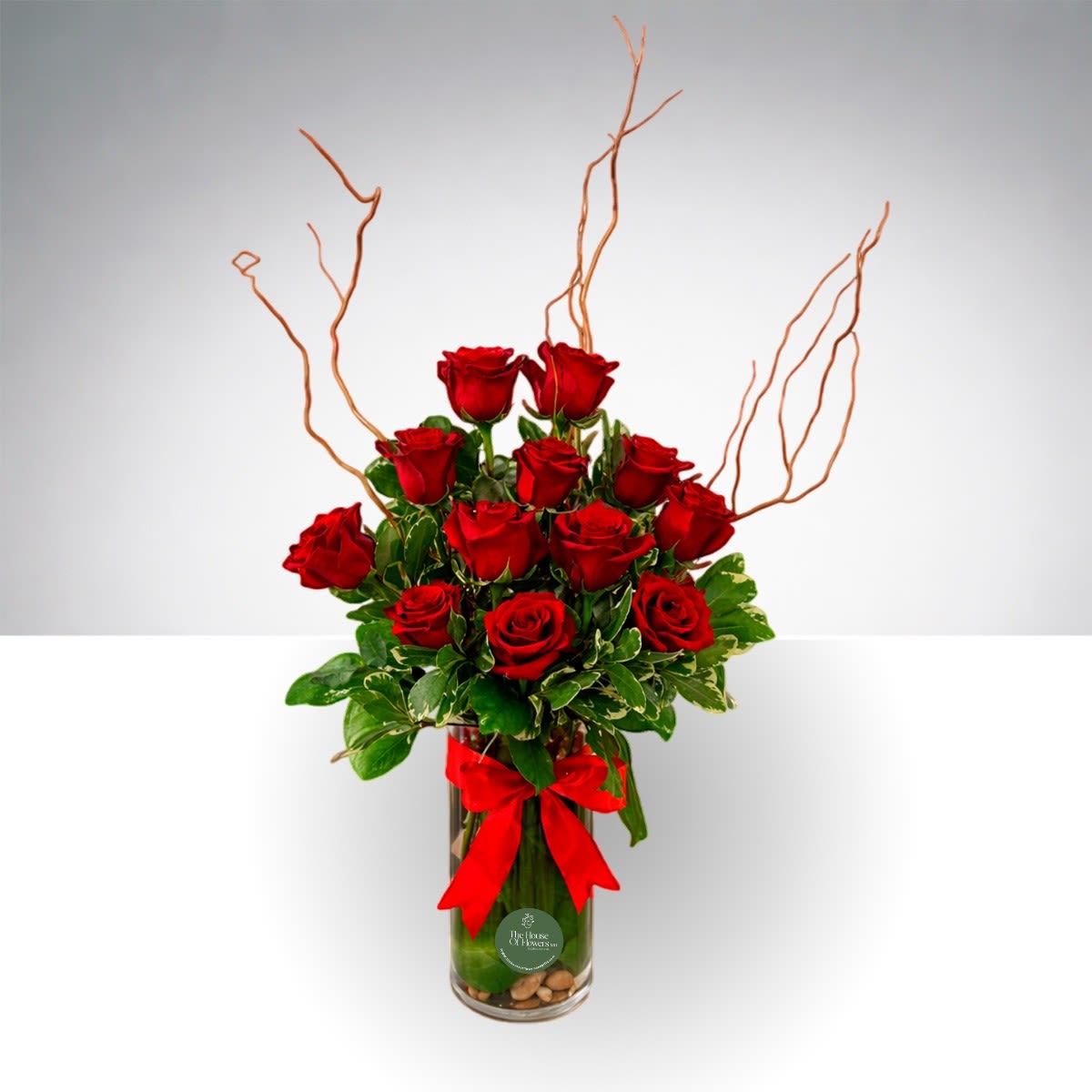 One &amp; Two Dozen Long Stemmed Roses by The House of Flowers - Classic 12 Rose Arrangement A timeless expression of elegance and love. This handcrafted design features 12 premium roses arranged in a modern glass vase with decorative stones at the base, fresh greenery, and natural accent branches for added height and dimension. Finished with an elegant ribbon detail.  Perfect for anniversaries, birthdays, romantic gestures, or simply to brighten someone’s day. Roses may vary in color based on availability and the client’s selection.   Fresh foliage  Decorative stones  Premium roses 