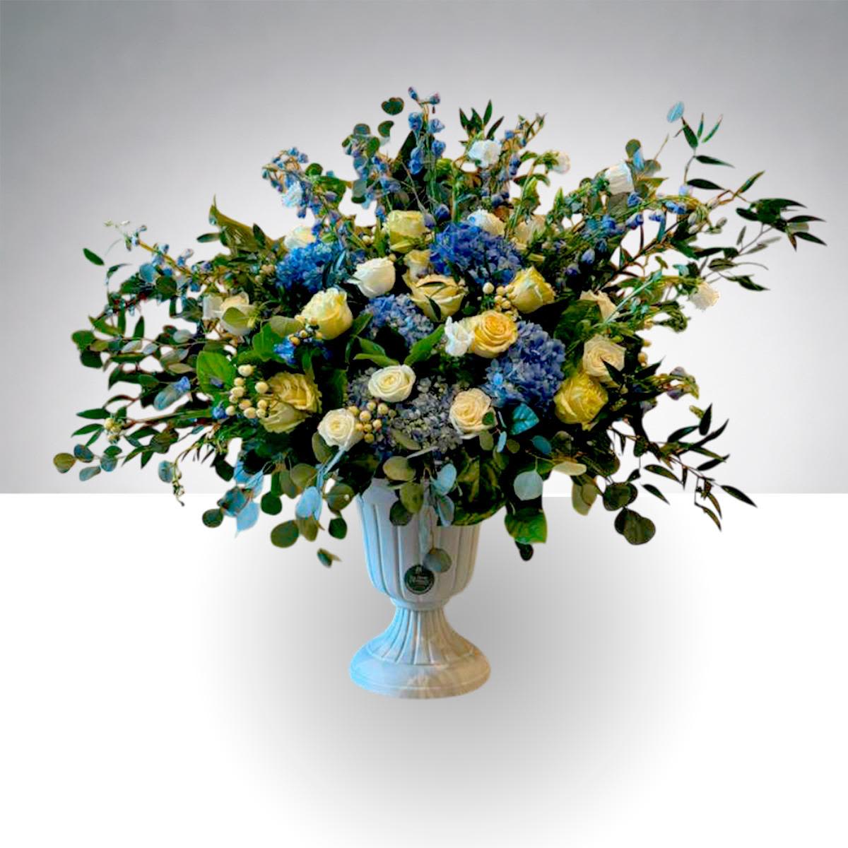 Grand Garden Elegance - This large and elegant floral arrangement, approximately 40 inches tall, is designed to make a beautiful statement at grand openings, inaugurations, and special events.  Created in a refined garden style, it features a graceful combination of soft blue hydrangeas, cream roses, delicate delphinium, and lush seasonal greenery. Arranged in a classic pedestal vase, the design offers a full yet airy composition that brings natural elegance and sophistication to any space.  Its impressive height and balanced shape make it perfect for welcoming guests at entrances, enhancing event spaces, or decorating corporate celebrations, galleries, showrooms, and formal gatherings.  Color palettes can be customized upon request. Clients may choose different color combinations depending on flower availability. Final design and pricing may vary based on selected flowers and seasonal availability. 