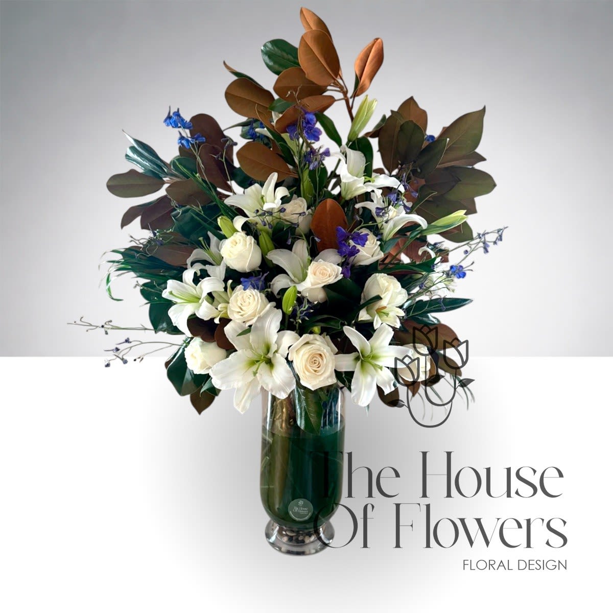 Magnolia Grand Elegance - A refined and elegant floral arrangement designed to make a beautiful statement in any space. This design features fresh white lilies, premium ivory roses, and lush magnolia foliage complemented with delicate blue accents.  Arranged in a tall clear glass vase with decorative stones, this piece offers impressive height and sophistication, making it perfect for home entrances, living rooms, office receptions, grand openings, or special occasions.  Approximate arrangement height: 30 inches, aprox. Vase height: 15-inch tall clear glass vase