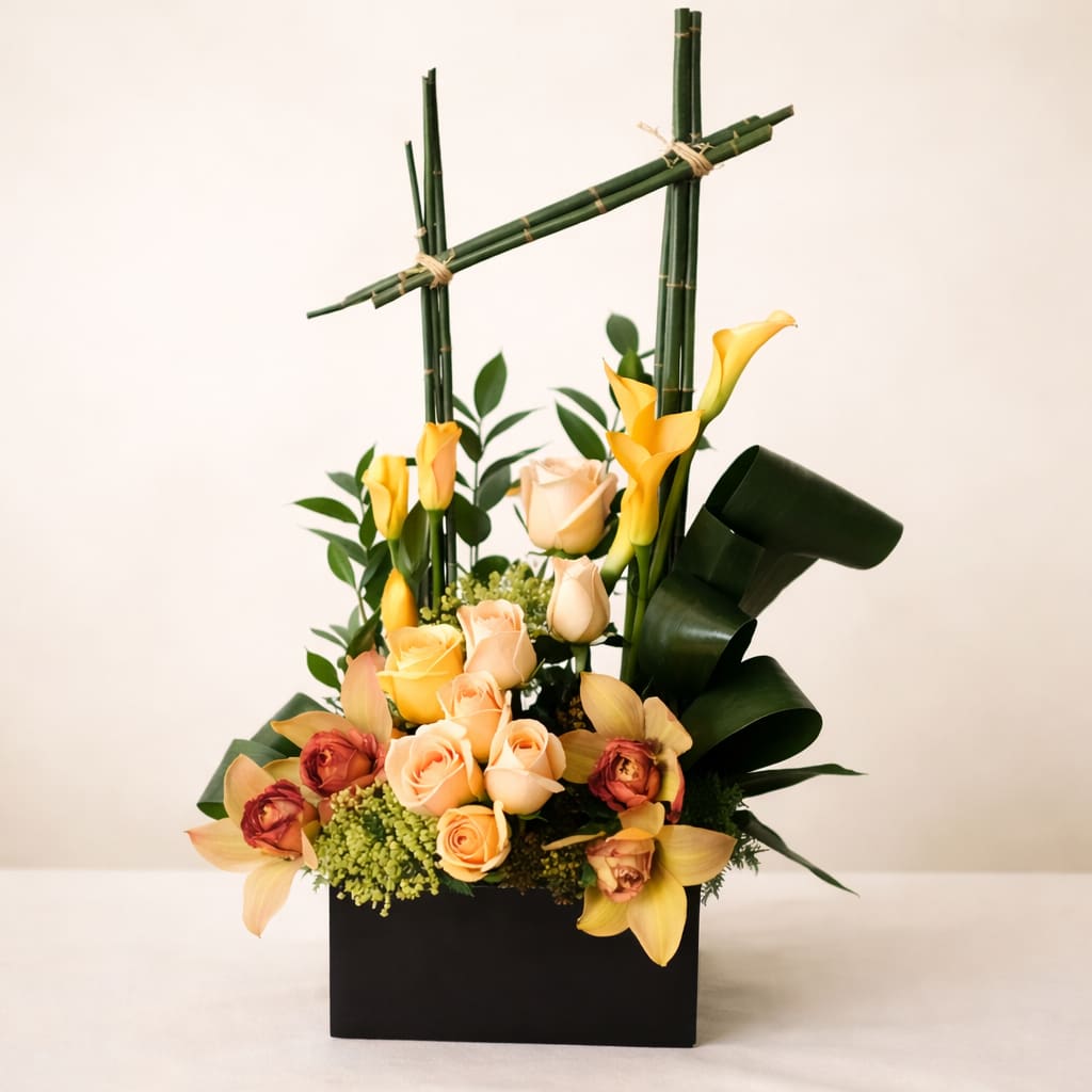 Scents of the Rainforest - Scents of the Rainforest is an elegant, sculptural arrangement inspired by the calm and freshness of a rainforest after rain, featuring soft peach and cream roses at its center, accented by vibrant yellow calla lilies, lush greenery, and natural textural elements, all framed by structured bamboo for an organic yet modern look, and presented in a sleek black or white square container that brings a sense of balance, sophistication, and natural luxury to any space. 