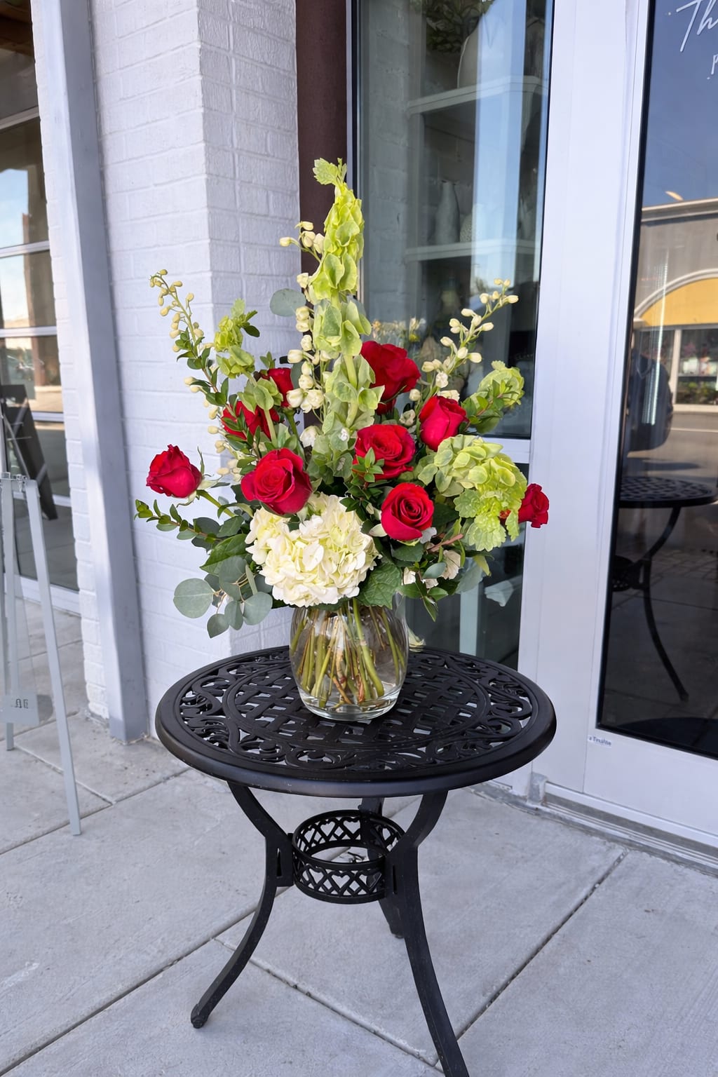 Crimson Garden Kiss - A timeless expression of love with a fresh, modern edge. This bouquet features a classic dozen velvety red roses nestled among soft hydrangea, sculptural Bells of Ireland, and layers of lush seasonal greens. Fragrant eucalyptus adds movement and a cool, aromatic finish, balancing the richness of the roses with natural elegance. Romantic, refined, and effortlessly beautiful—perfect for grand gestures or meaningful moments.