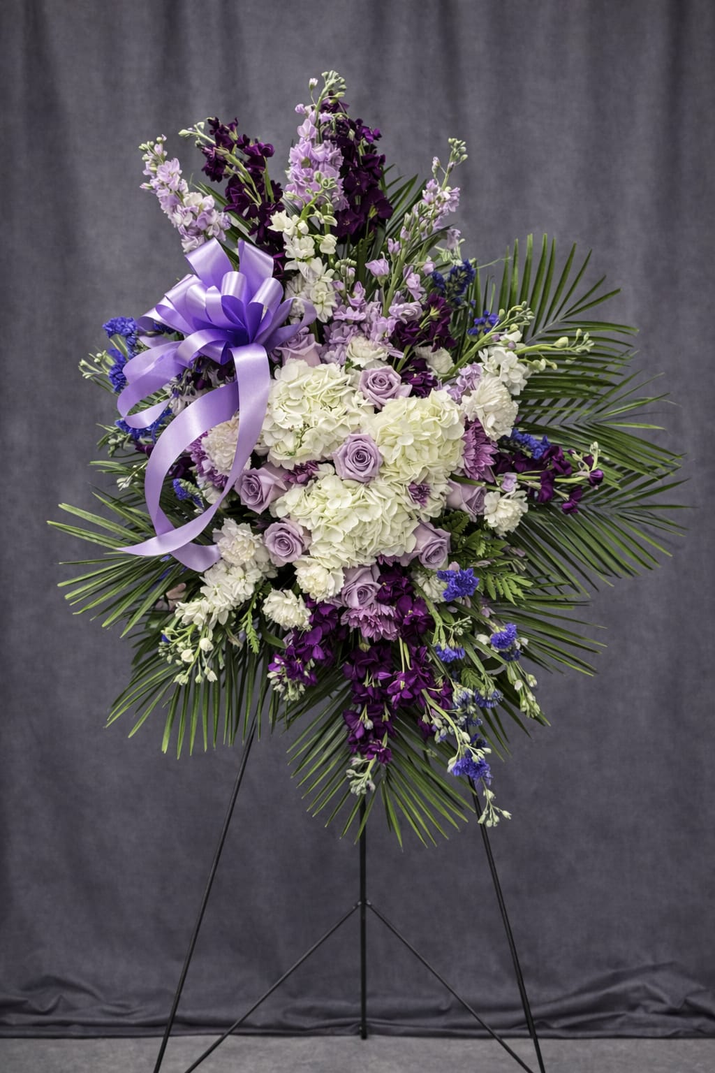 Lavender Remembrance - Lavender Remembrance is an elegant and uplifting standing tribute designed in soft shades of lavender, white, and deep purple. Lush white hydrangea forms the heart of the arrangement, accented with delicate lavender roses, rich purple stock, statice, and fresh greenery for height and graceful movement. A satin lavender bow adds a refined finishing touch.  The soothing color palette symbolizes peace, dignity, and heartfelt remembrance, making this spray a beautiful expression of love and honor. Displayed on a standing easel, Lavender Remembrance offers comfort and quiet strength during services, memorials, or celebrations of life.