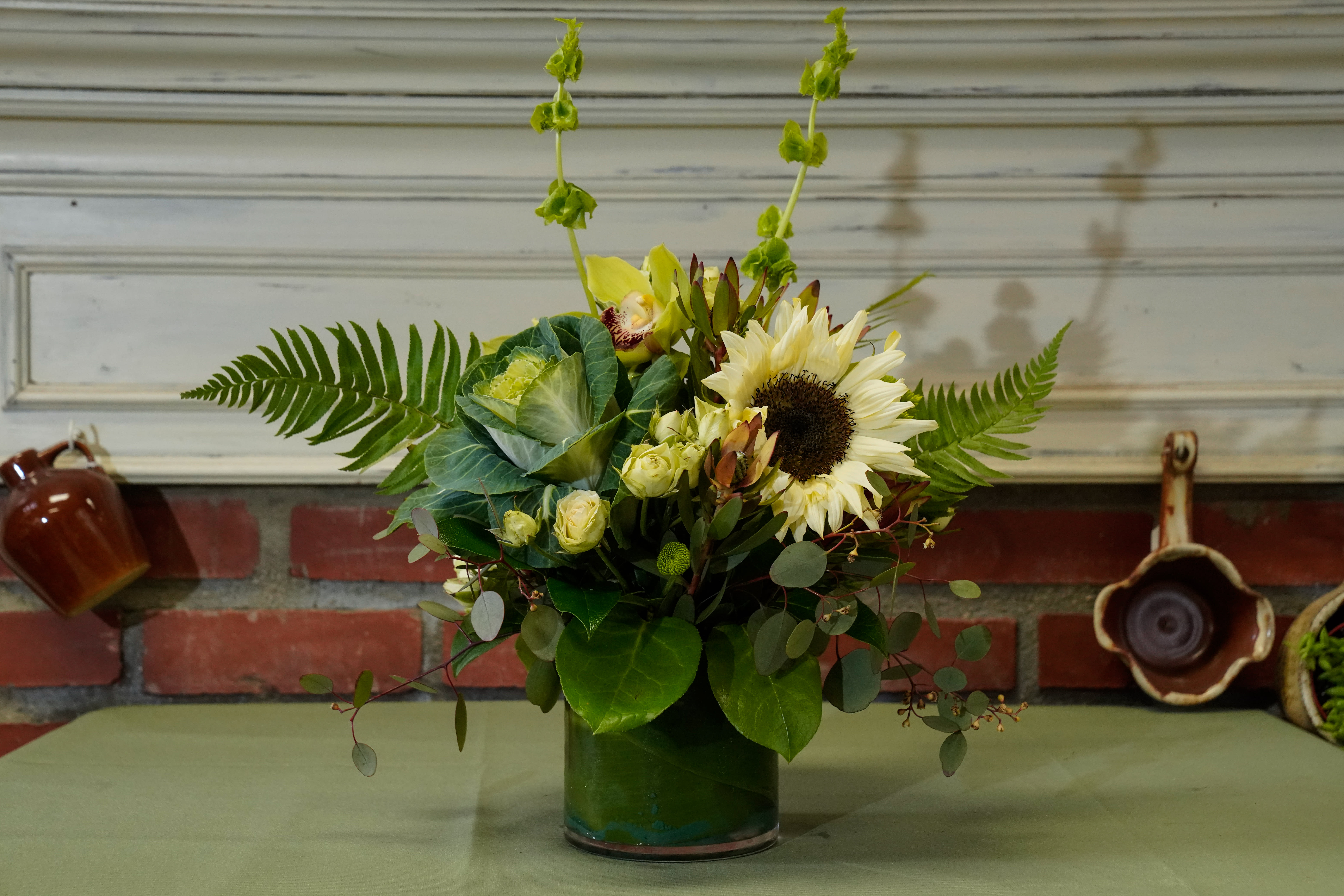 Lush Green Hillside - Type of Flowers: Bells of Ireland, Kale, Sunflowers, Spray Roses, Button Mums, Safari Hypericum Berries, and more in a short vase wrapped with a green leaf. Availability: All year round Design View: All Facing View Photo shown: Standard 