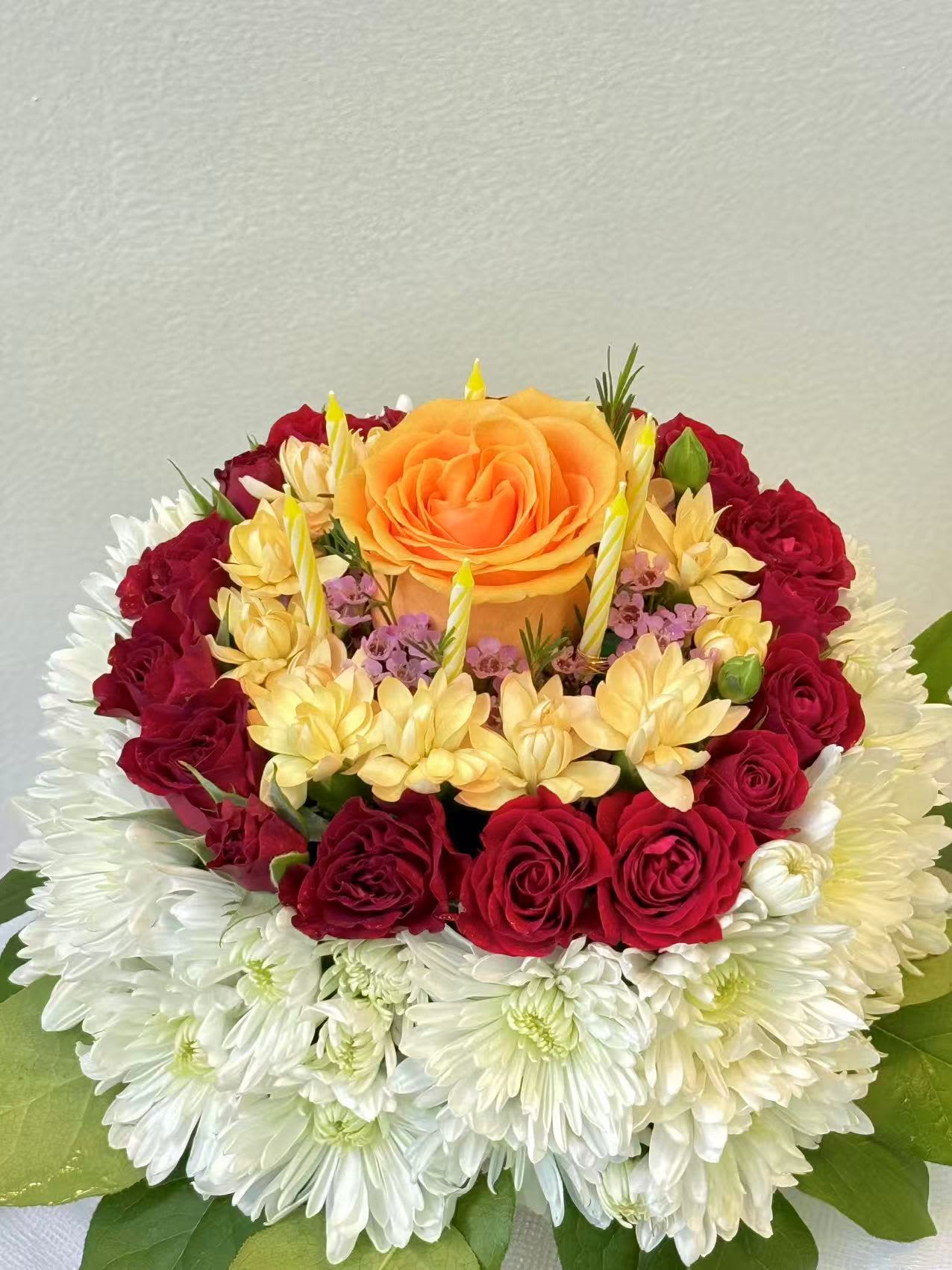 Birthday Bloom Cake - A delightful floral arrangement designed to resemble a birthday cake, featuring roses, chrysanthemums, and seasonal blooms topped with festive candles. This cheerful design is the perfect way to celebrate birthdays and special moments with flowers instead of cake.