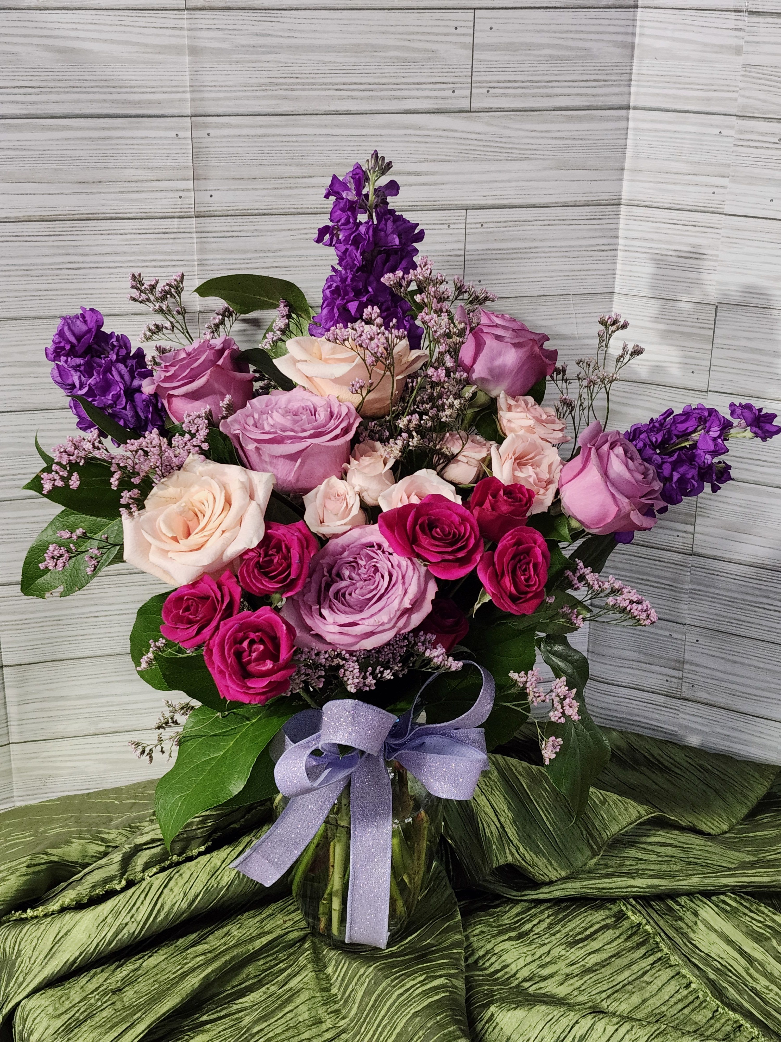 Designer's Choice Valentine Variety Mix Flowers - One sided,  Mix flowers of different Valentine colors of pinks, purple, lavender standard is 6 different roses and stock and filler in a Valentine color vase and matching bow