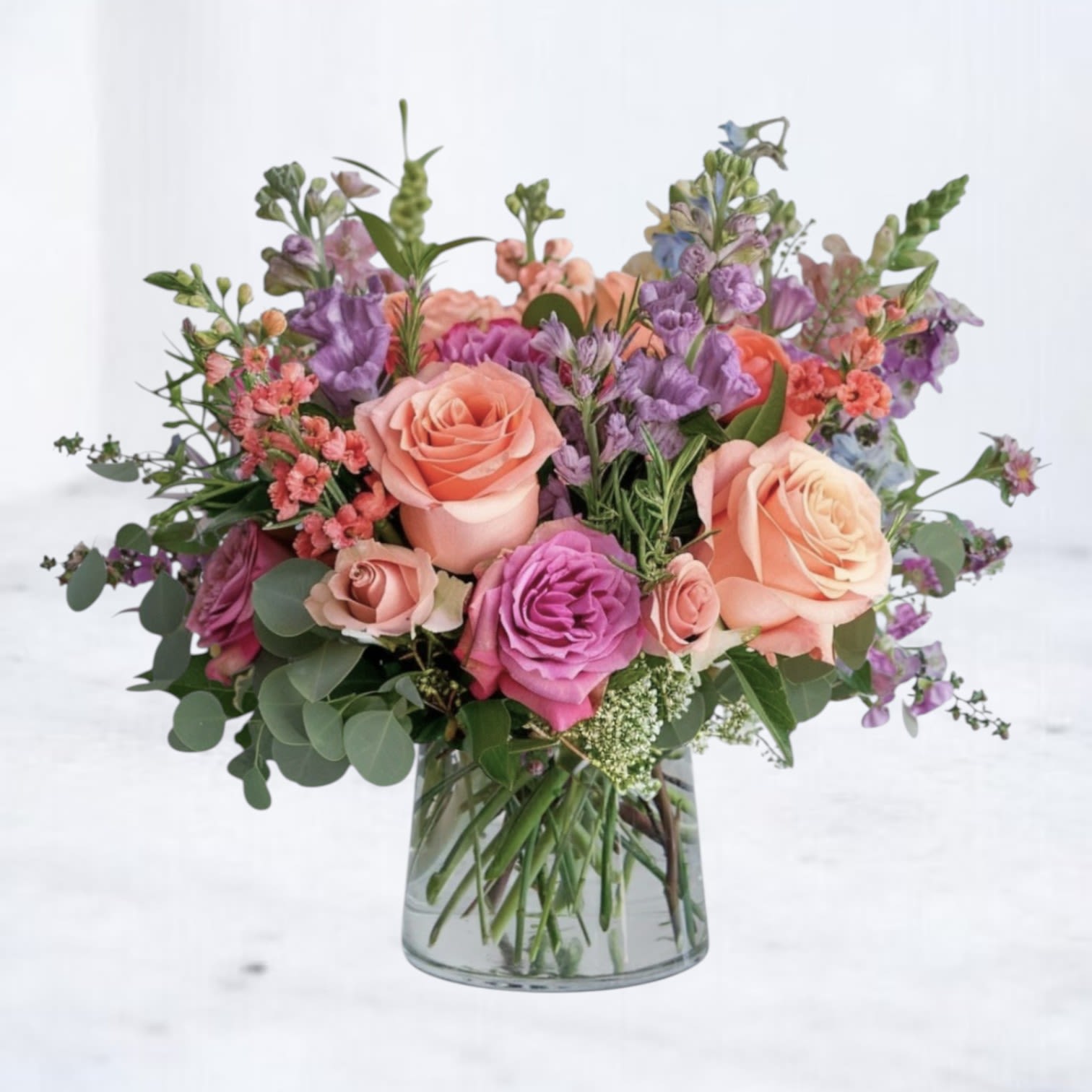 Soraya Charm - Bright, cheerful, and artfully arranged, the Soraya Charm bouquet brings a burst of modern garden beauty to any space. Featuring a harmonious blend of vibrant coral roses, soft peach blooms, vivid fuchsia spray roses, delicate purple stock, and airy accents of seeded eucalyptus, this lush arrangement feels freshly gathered from a blooming meadow. Its loose, airy design and layered textures make it the perfect gift for birthdays, anniversaries, thank-you gestures, or simply brightening someone’s day. Designed with a modern twist on classic floral beauty, the Soraya Charm is hand-delivered in a stylish glass vase for a polished, ready-to-enjoy display. 