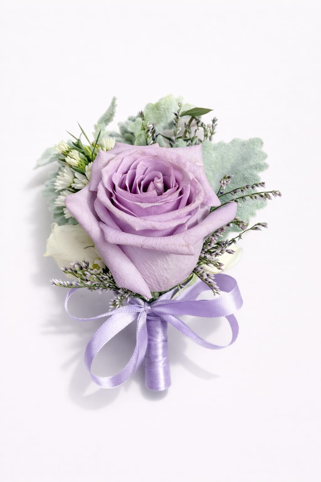 Lavender Whisper Boutonniere - Delicate, elegant, and full of charm, this beautiful boutonniere features a soft lavender rose accented with fresh greenery and subtle filler blooms. Finished with a satin ribbon wrap, this refined piece adds a touch of romance and sophistication to any special occasion.  The gentle lavender tones symbolize grace, admiration, and love, making it a perfect detail for weddings, proms, quinceañeras, or formal celebrations.  Simple, timeless, and effortlessly elegant.