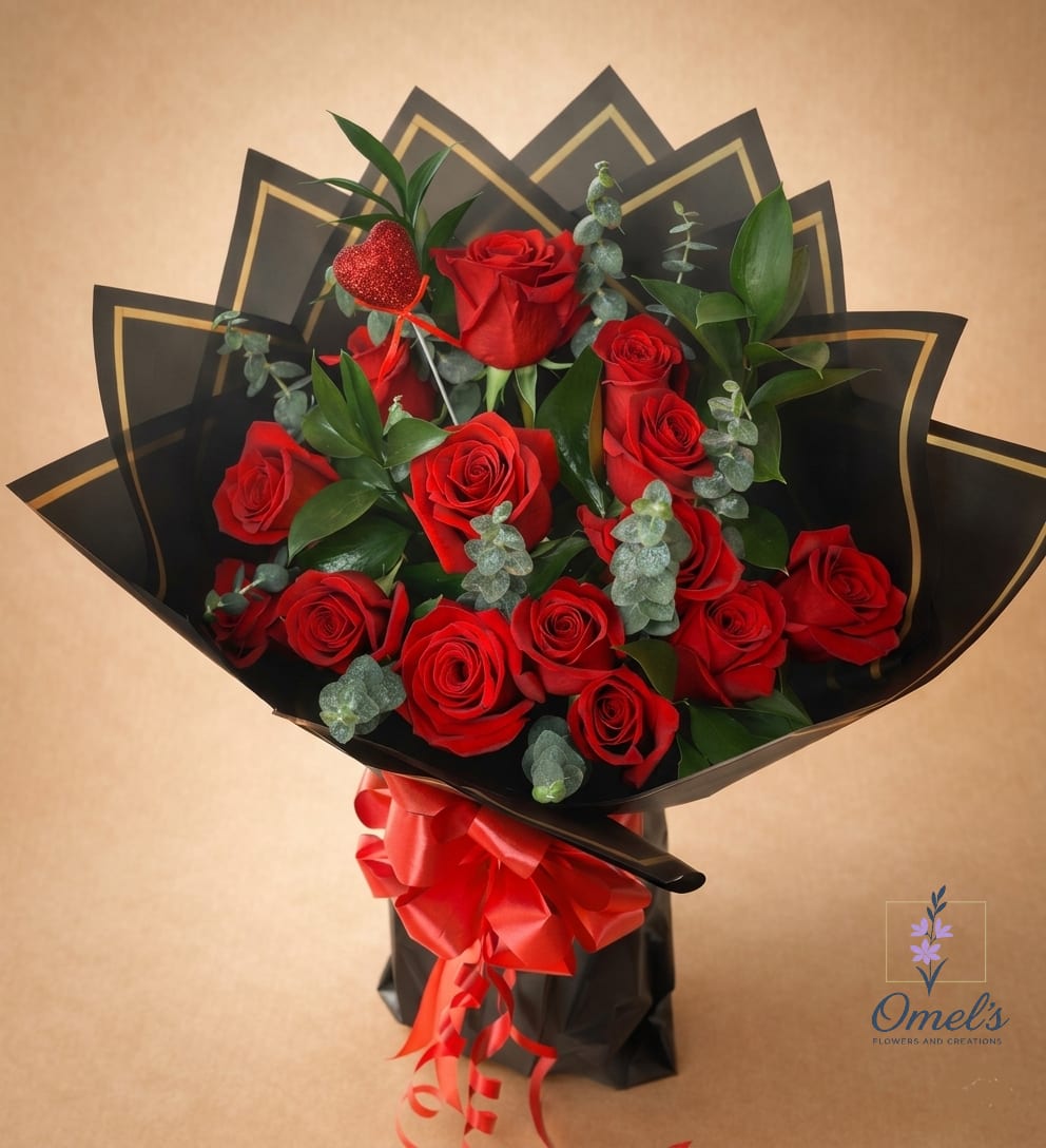 Crimson Bouquet  - A captivating blend of classic romance and modern drama. This stunning bouquet features a vibrant collection of premium, rich red roses, their velvety petals contrasting beautifully with the cool, silvery-green tones of fresh eucalyptus sprigs and the deep, glossy leaves of ruscus greenery. The arrangement is expertly wrapped in striking, origami-folded black paper, edged with a sophisticated gold border, creating a fan-like frame that draws the eye. To complete the look, a large, brilliant red satin bow rests at the base, and a sparkling red heart accent adds a charming touch of passion. Presented in a sleek black, bag-style container, this is more than a gift; it is an experience of pure elegance and luxury. Perfect for celebrating a milestone anniversary, a formal romantic gesture, a sophisticated Valentine's Day surprise, or to simply make an unforgettable impression on someone special.