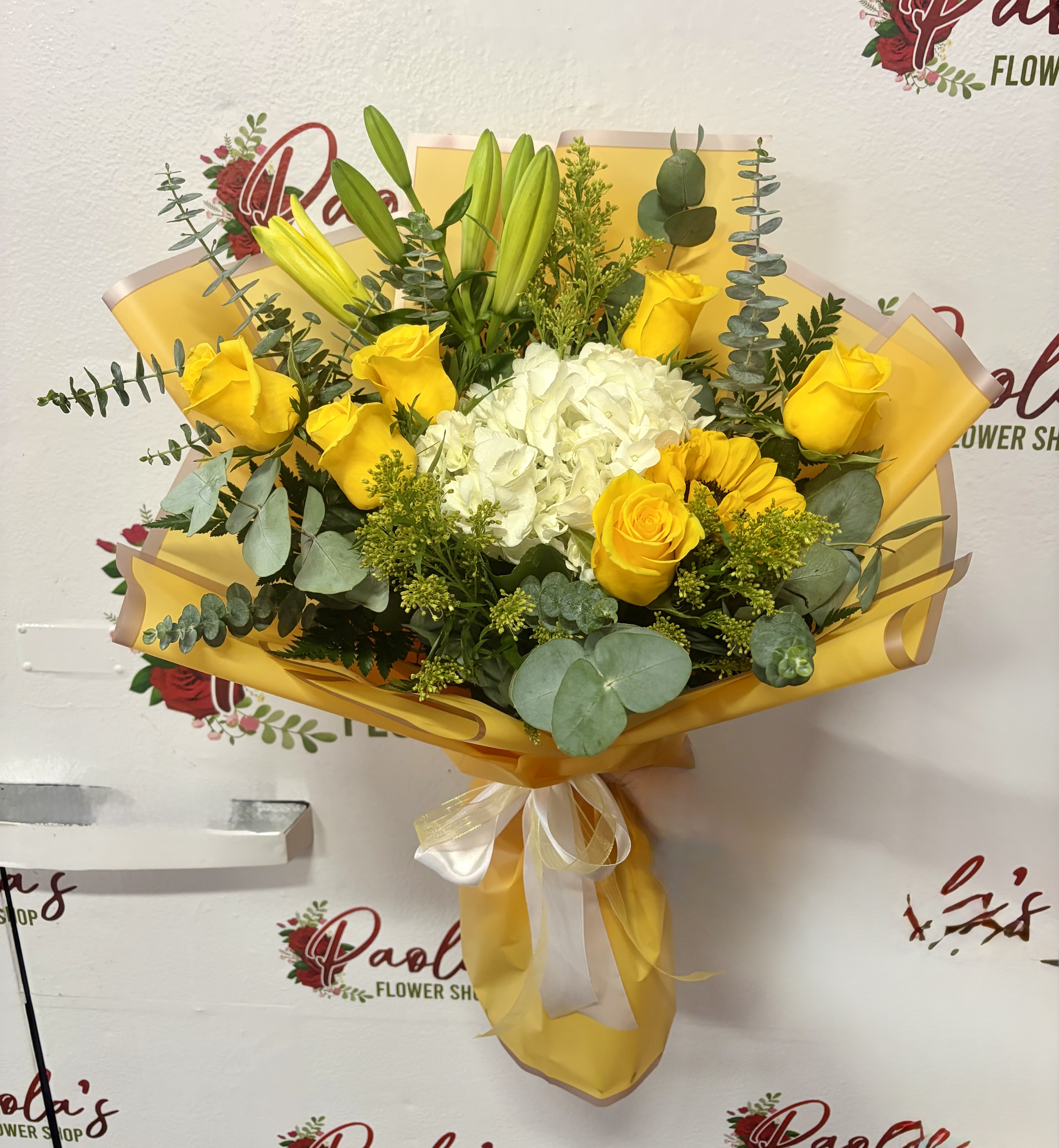 Golden Sunshine Bouquet - Brighten someone’s day with our Golden Sunshine Bouquet, a cheerful arrangement designed to bring warmth and happiness. This hand-wrapped bouquet features vibrant yellow roses, elegant lilies, a soft white hydrangea centerpiece, seasonal yellow accents, and fresh eucalyptus greenery. Wrapped in premium yellow floral paper and finished with a delicate ribbon, this bouquet is perfect for birthdays, celebrations, thank-you gifts, or simply sending a little sunshine to someone special.  Flowers may arrive in bud form to ensure longer freshness and bloom beautifully over the next few days.