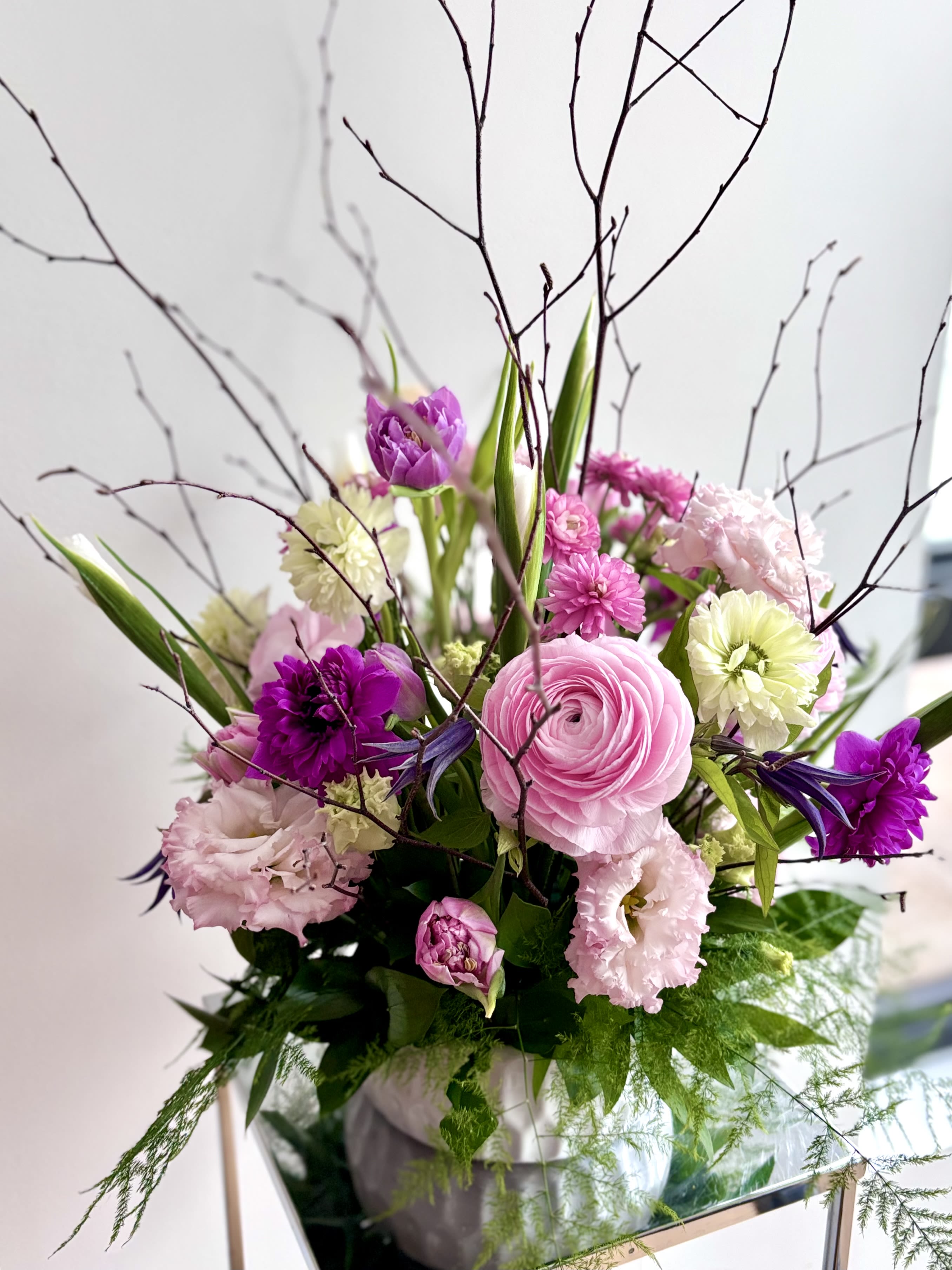 Easter Morning Garden - A fresh flower centerpiece for Easter with a rich variety of premium spring flowers in the color palette of pinks, fuchsias, purples and whites. The selection of flowers may vary, but rest assured that Michaela will work with a beautiful variety of flowers such as full star anemones, lisianthus, clematis, iris, tulips and ranunculus. Twigs and fern greenery will complete the unique creation for your table. The container may vary based on what we have available in the studio - usually white ceramic vase.