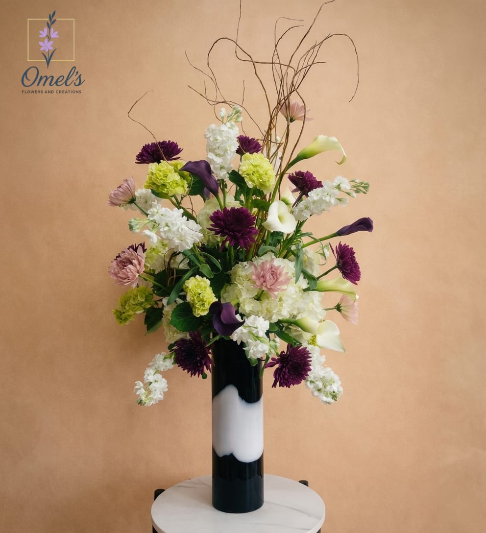 Wild Elegance - A sophisticated design full of movement that features wild beauty with a contemporary style. The delicate white Calla Lilies accompanied by deep violet-purple and lavander Mums mixed with green Hydrangeas and white Stock. The accented twigs create a modern, artistic and tall silhouette.  A tall, distinctive cylindrical ceramic vase featuring a matte midnight-black glaze with a dramatic, central band of glossy bone-white glaze.   Ideal Occasion to Use: This arrangement's sophisticated color palette and striking, graphic design make it ideal for a modern event or high-end setting. Perfect for a corporate centerpiece, a modern wedding reception, a chic anniversary celebration, or a sophisticated sympathy offering. It is a bold, modern choice that guarantees to make an elegant statement.