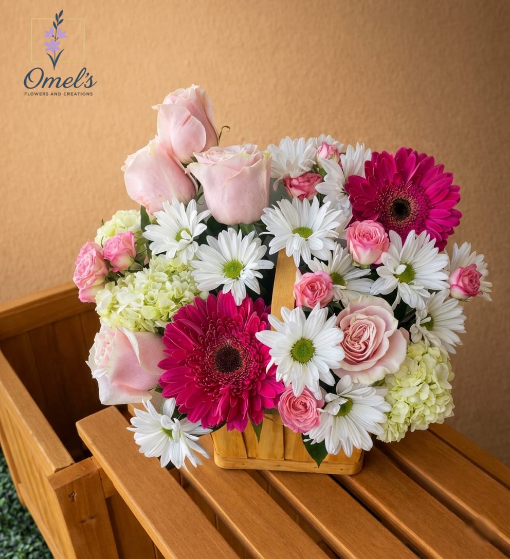 Blush Garden Basket - Bring the charm of a sun-drenched cottage garden indoors with this exquisite, abundant arrangement, hand-crafted to celebrate the softest hues of hot pink Gerbera daisies that provide a joyful focal point, contrasted by the delicate, multipetal form of elegant blush-pink roses. Clusters of white cushion poms and soft-chartreuse hydrangeas add incredible texture and depth, all nestled within a charming, light-wood wicker basket with a classic central handle.  Perfect for Mother's Day; to say "I love you" to a partner; a sophisticated "Get Well Soon" gesture, or as a timeless centerpiece for a bridal or baby shower.
