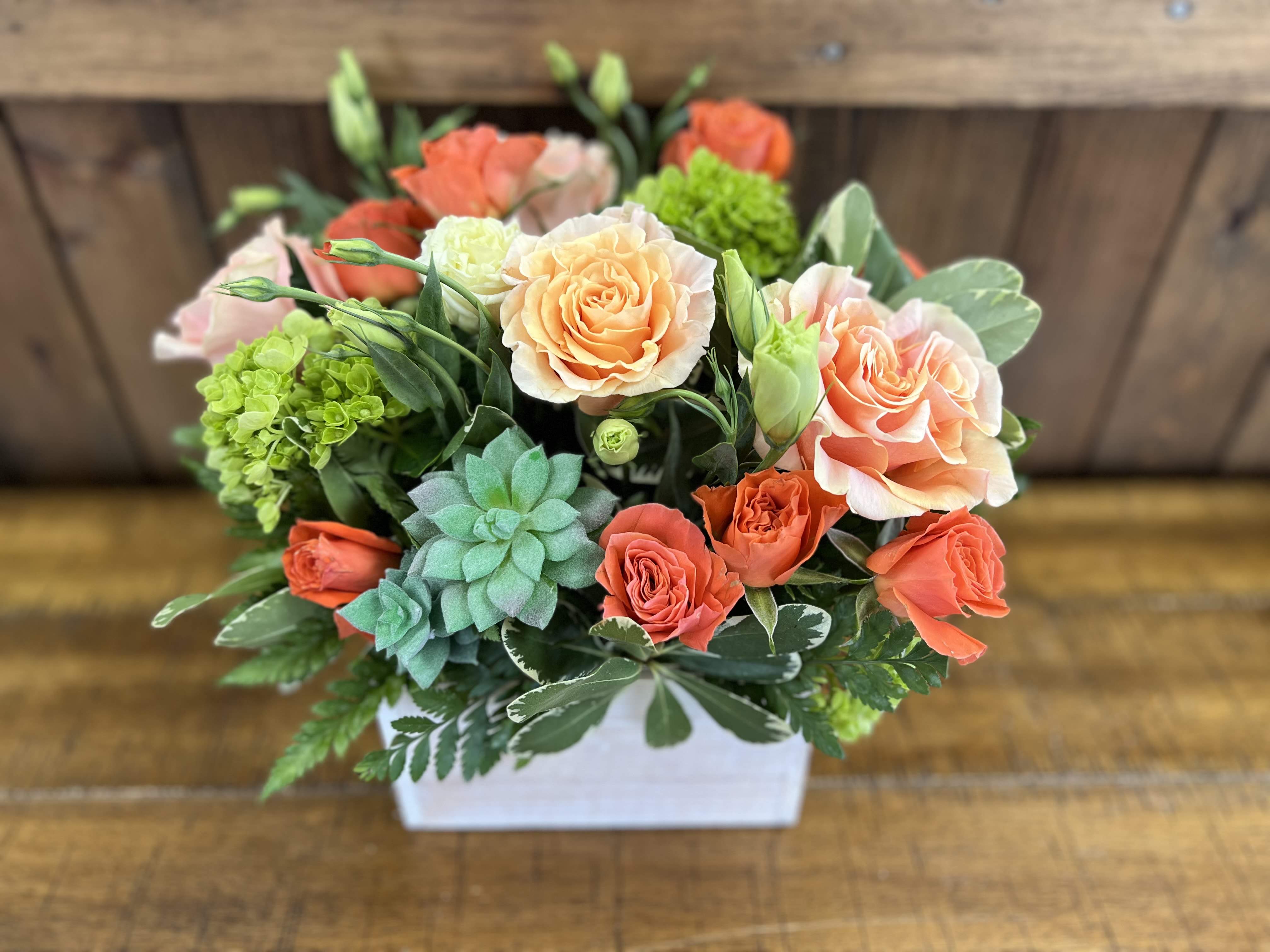 Peaches - A stunning arrangement featuring soft peach roses nestled among lush green hydrangea and vibrant orange carnations. This beautiful mix is artfully arranged in a rustic white-washed box, enhancing the overall charm and vintage feel.