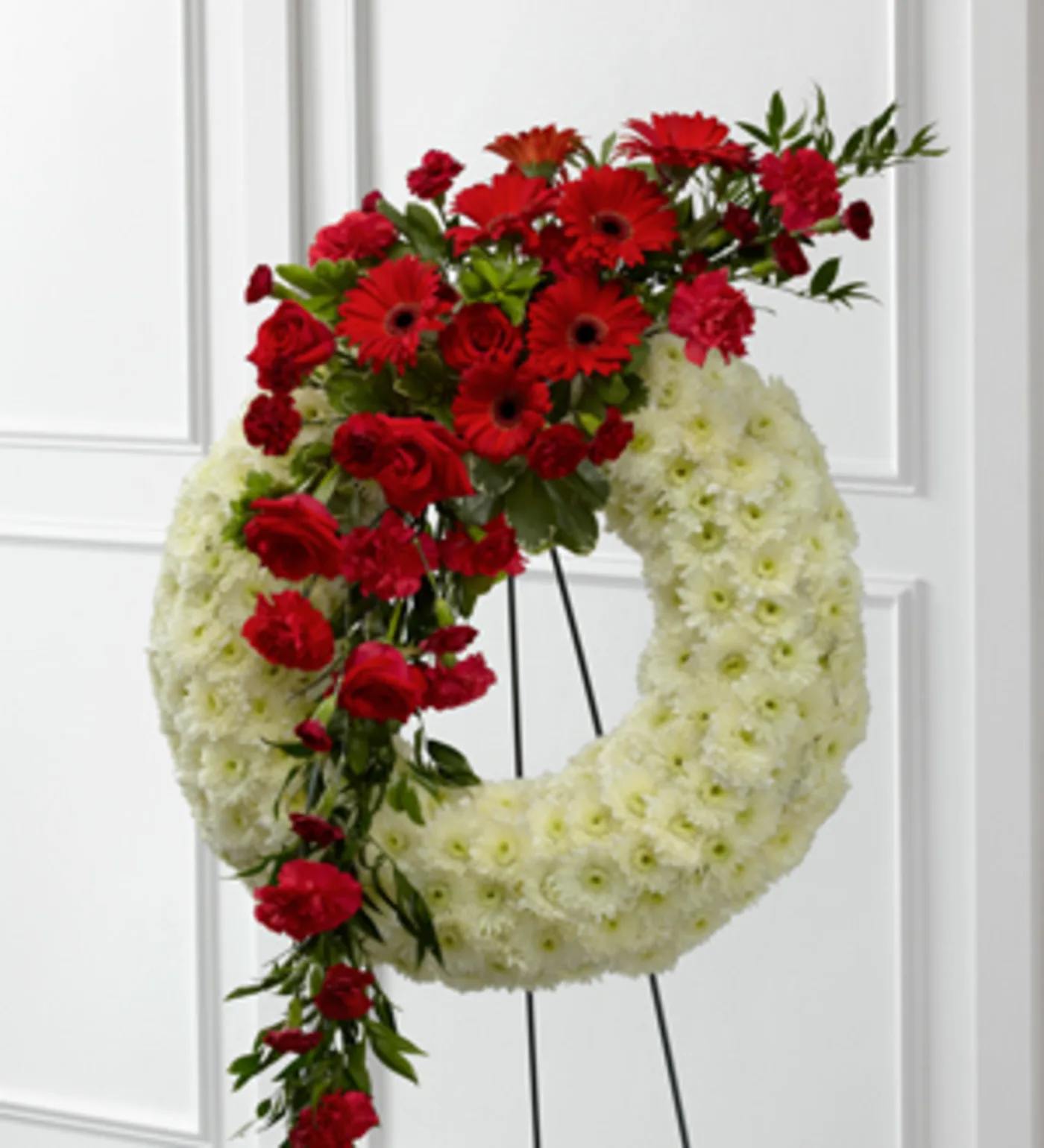 Graceful Tribute Wreath (Luxury line) - Graceful Tribute™ Wreath creates a stunning presentation of love's serenity for their final farewell service. Red roses, carnations and gerbera daisies are accented with burgundy mini carnations and a variety of lush greens to create a sweeping arrangement that glides across the face of a snow white wreath consisting of chrysanthemums making this a remembrance for one who has touched your life like none other. Displayed on a wire easel.