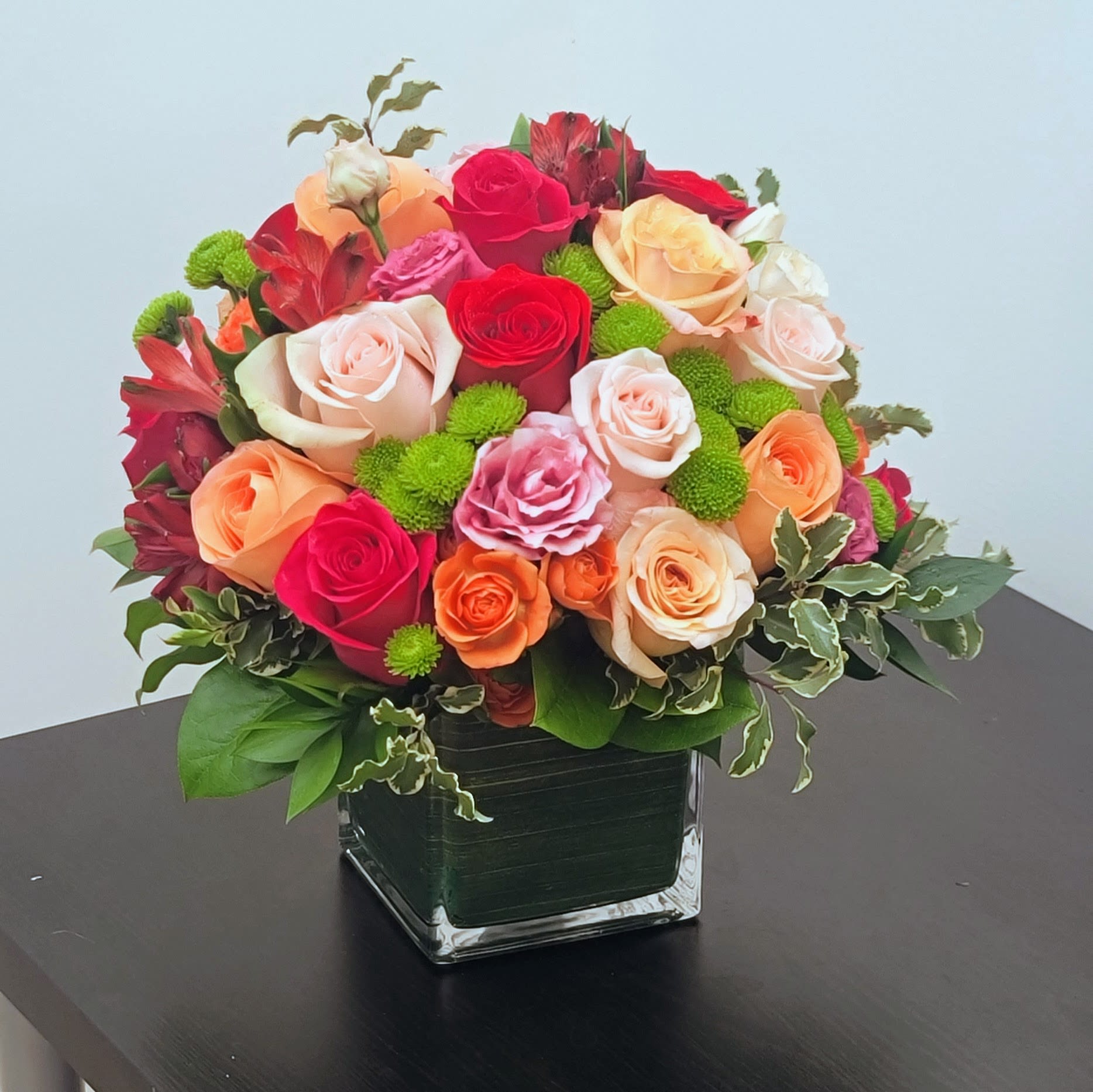 Kaleidoscope Bouquet - Experience the vibrant spirit of a garden with this exquisite bouquet. A mesmerizing blend of roses in hues of pink, orange, and yellow, adorned with lush greenery and delicate pink blossoms, gracefully arranged in a modern glass vase.  This captivating ensemble is a visual delight, ideal for every celebration.  Approximately 14" H  and 14" W.