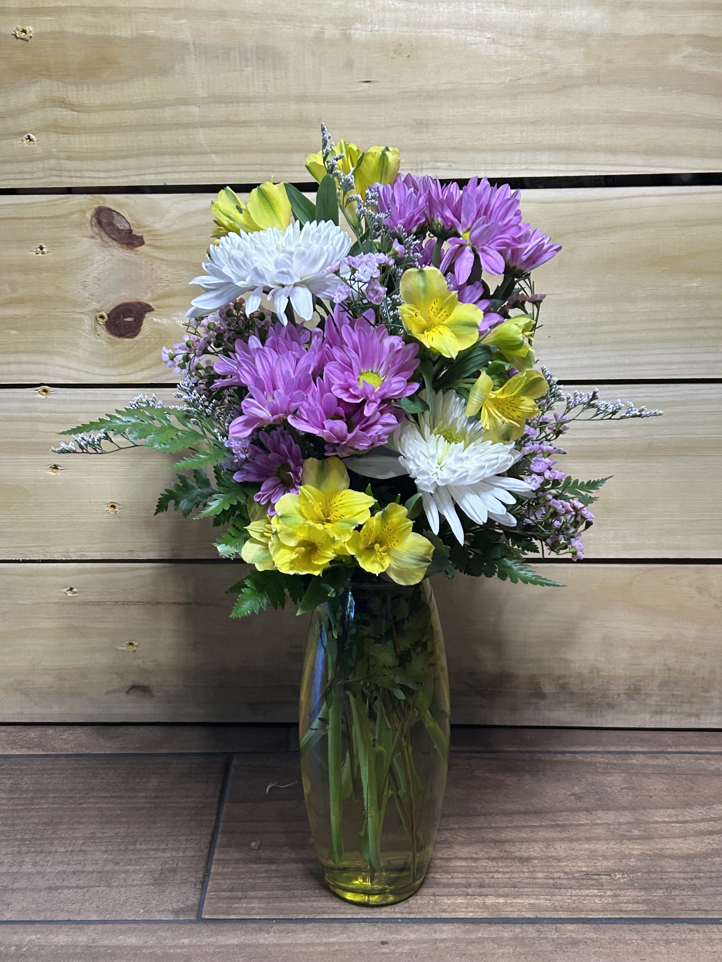 Sunshine Garden Bouquet - Bring a little sunshine indoors with the Sunshine Garden Bouquet. This cheerful arrangement features vibrant yellow alstroemeria, soft lavender daisies, and crisp white mums nestled among delicate waxflower and fresh greenery. Designed in a clear glass vase, its bright colors and garden-fresh style make it the perfect pick-me-up for birthdays, thank-yous, or simply brightening someone’s day. A joyful blend of blooms that feels like a warm spring morning.