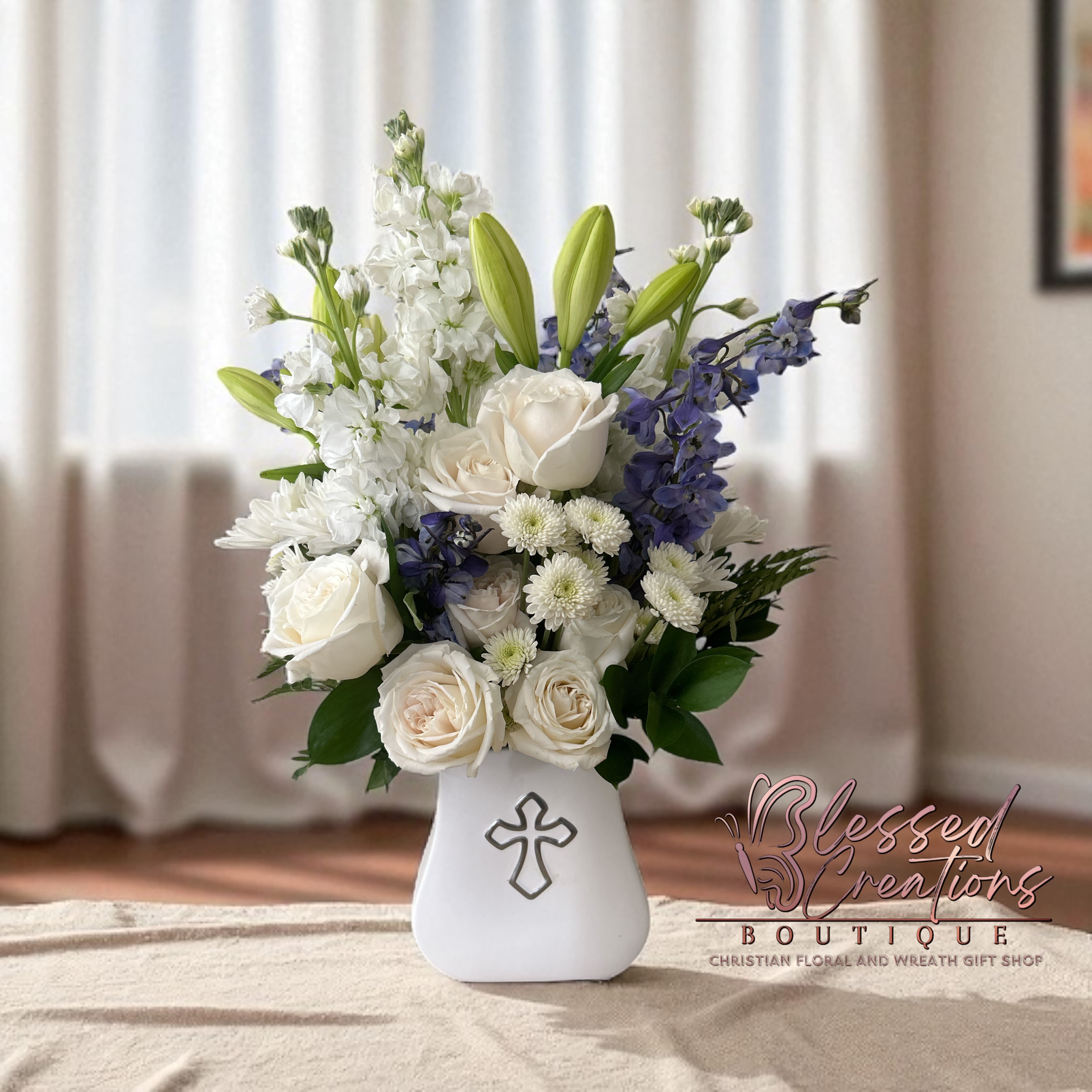Heavenly Peace Sympathy Bouquet  - The Heavenly Peace Sympathy Bouquet is a serene and elegant display designed to bring comfort during times of loss. This arrangement features pure white roses, white stock, white lilies, and delicate white filler flowers, accented with blue delphinium for a touch of calming color. Nestled among lush greenery, the blooms are arranged in a graceful white vase adorned with a cross, symbolizing faith, hope, and everlasting peace. A thoughtful gift to honor a loved one and express heartfelt condolences.