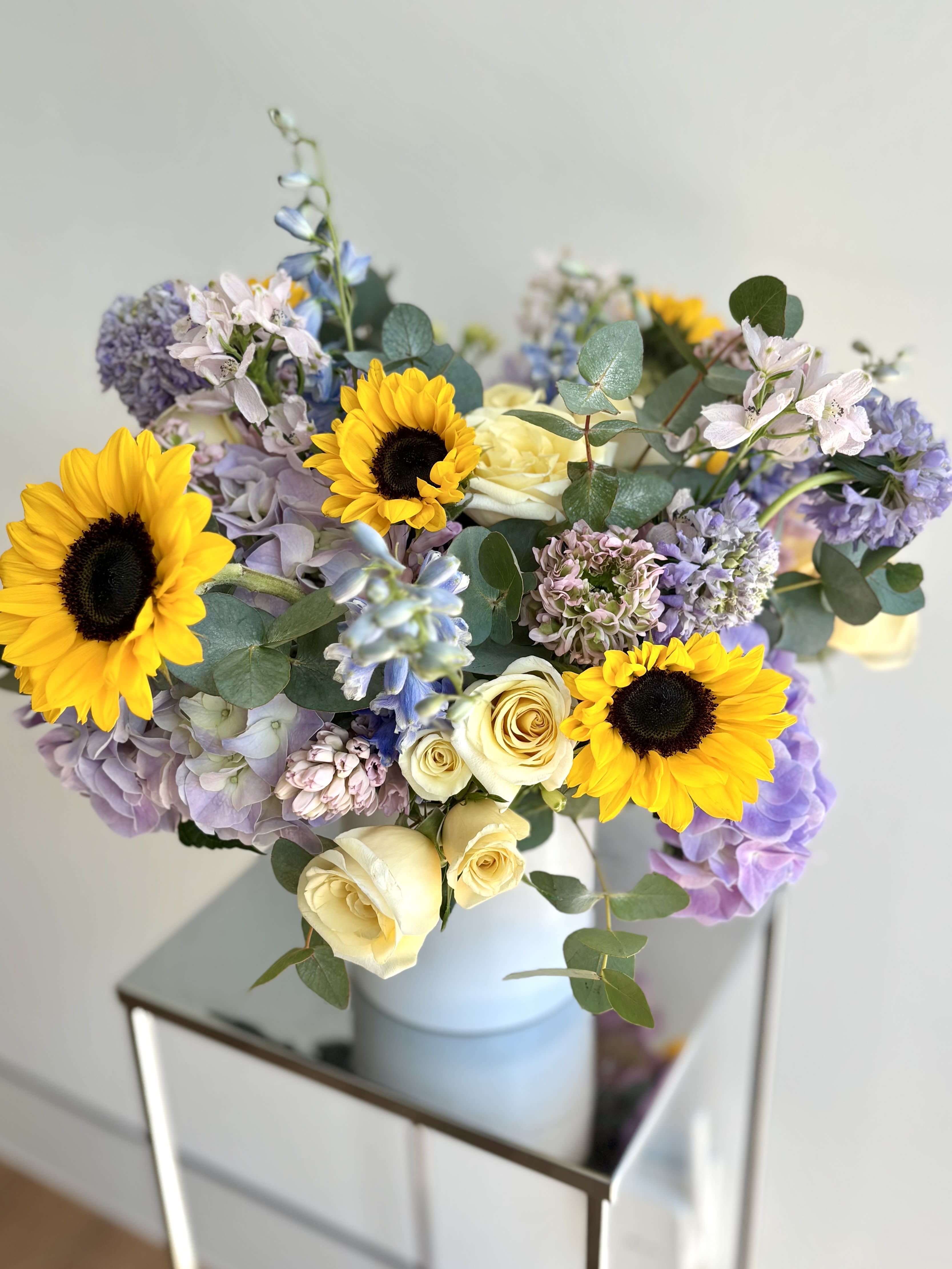 Blue Skies &amp; Sunflowers - A lovely flower arrangement with soft purple and sky blue flowers, creamy yellow, with a pop of sunflowers in a white 5inch cylinder vase. In addition to sunflowers we usually mix in hydrangeas, stock, spray roses, delphinium, hyacinths, and additional premium flowers and eucalyptus. The selection of flowers may vary based on the availability but our designer always selects the most fresh flowers we have in the studio.