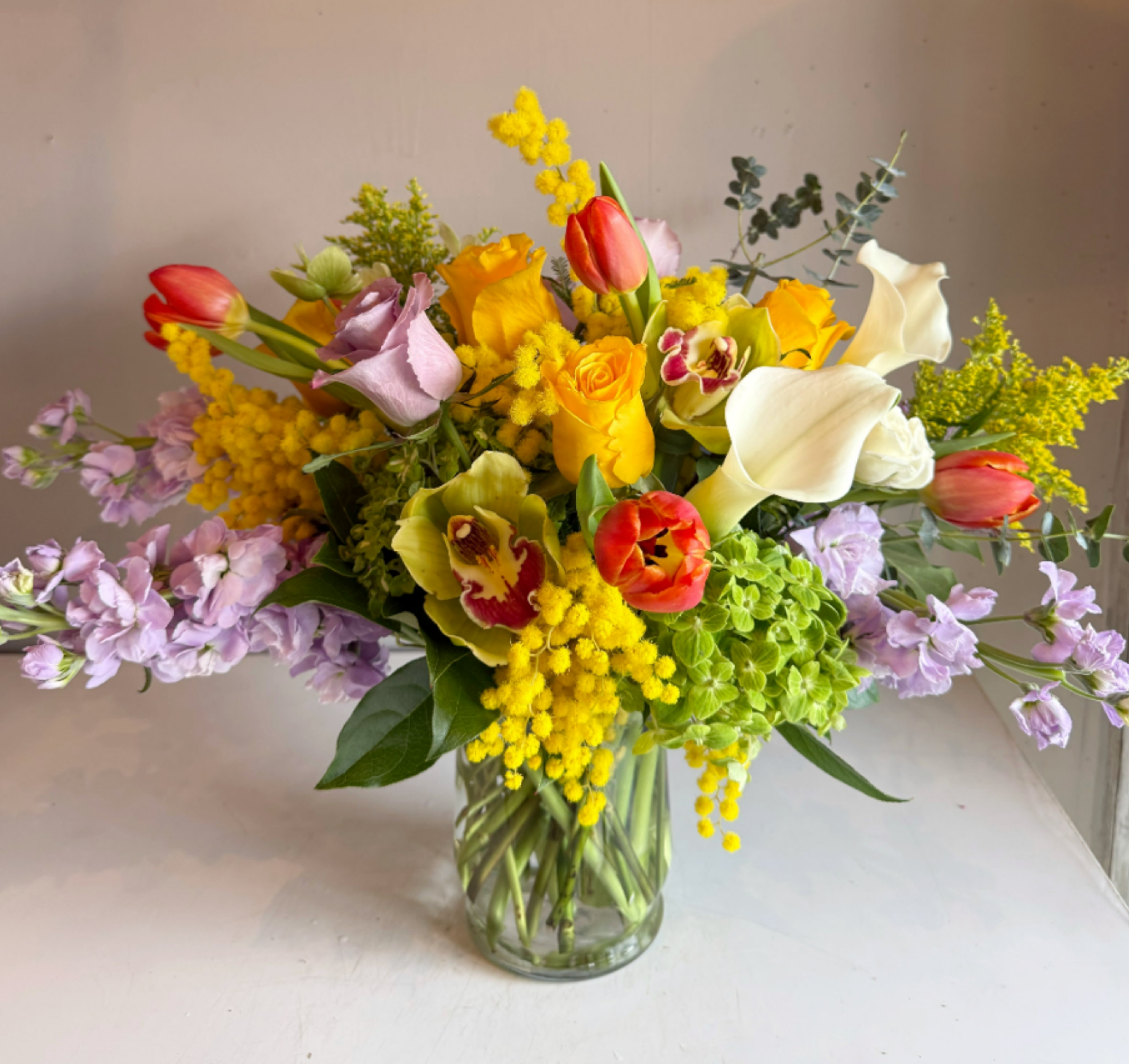 Spring Melody - A cheerful arrangement consisting of bright Spring flowers such as Brighton roses, lavender stock, cymbidium orchids, Acacia mimosas, and tulips.