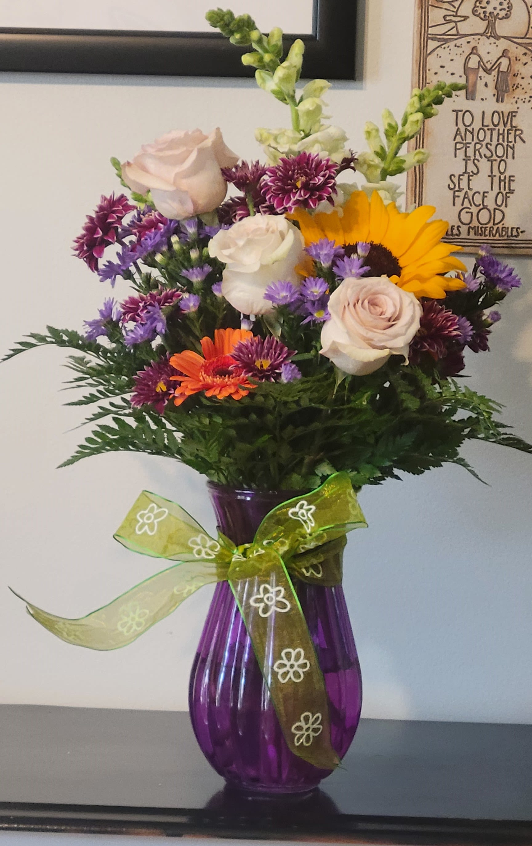 Sunshine and Smiles by Dixie's Flowers - "A bright and cheerful mix of sunflowers, roses, daisies, and snapdragons, arranged in a purple vase with a playful green bow—perfect for sending a little sunshine someone's way!"