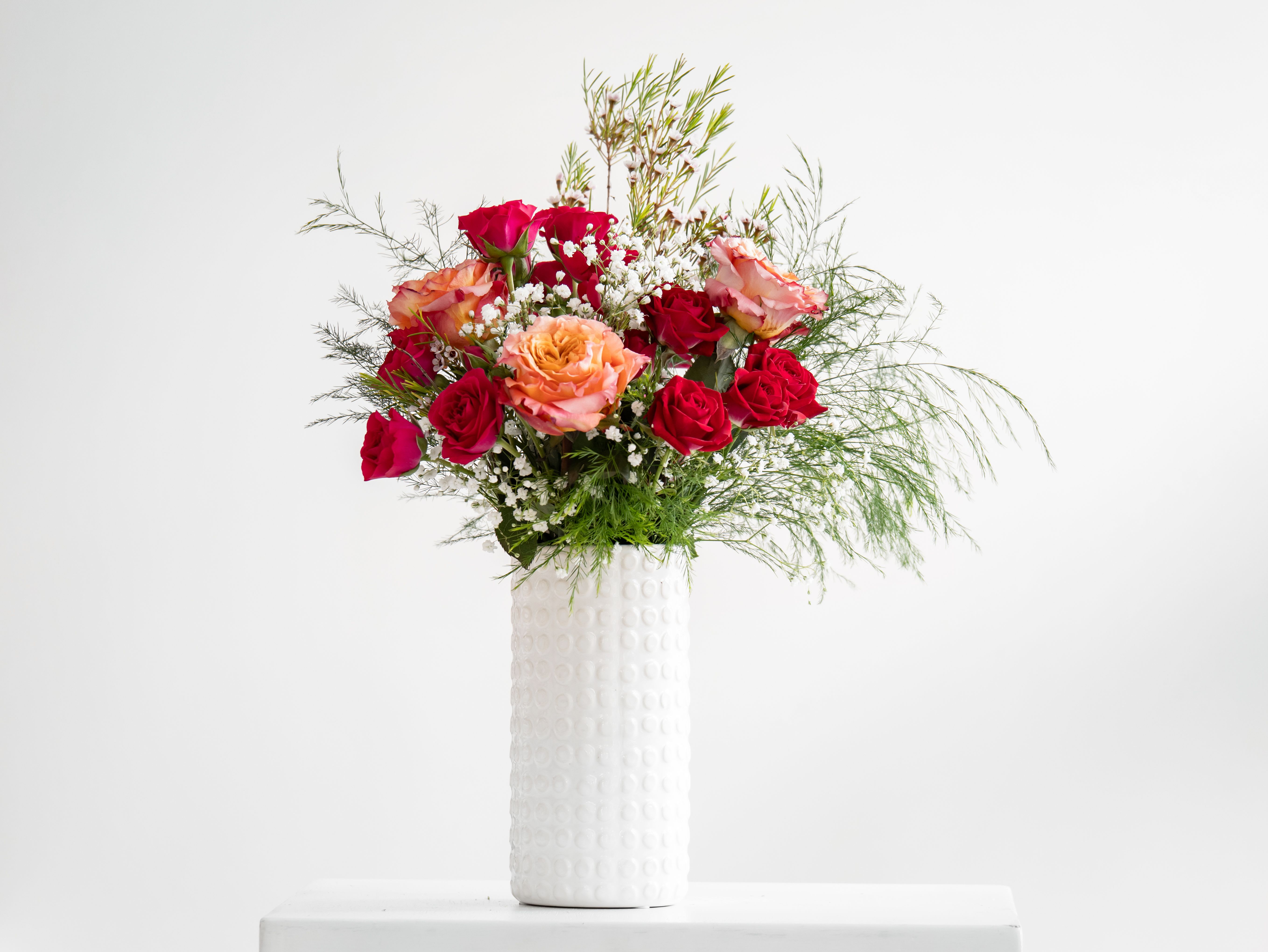 Tall arrangement of red and peach roses with white filler flowers in a textured white vase