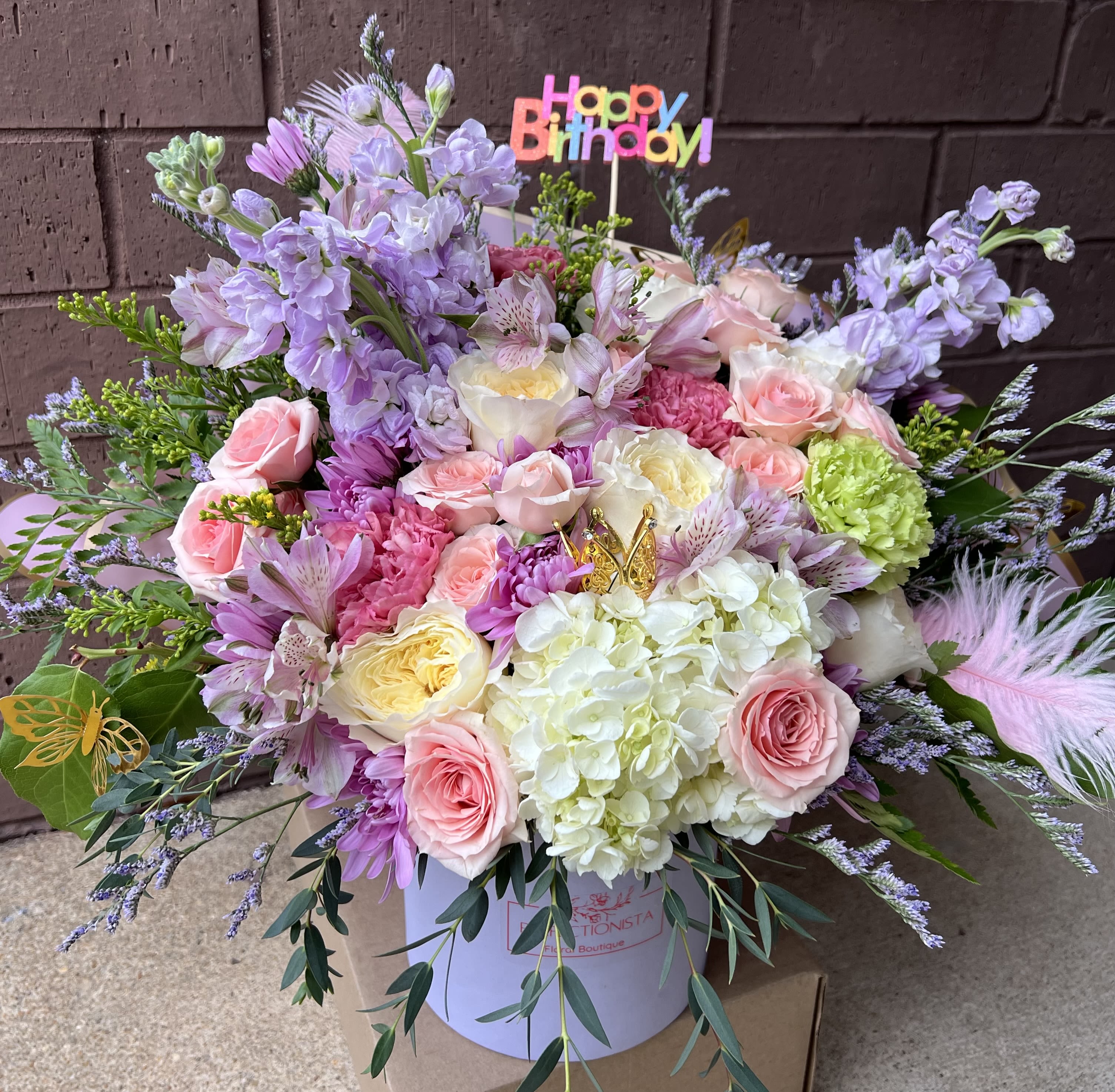 Lavender Dreams - A dreamy pastel arrangement designed to make any birthday unforgettable. This lush hatbox features a soft and romantic blend of roses, hydrangeas, spray roses, alstroemeria, stock, and seasonal blooms in delicate shades of blush pink, cream, lavender, and mint green. Accented with airy greenery, whimsical feathers, and a butterfly touch, it exudes elegance and charm.  Presented in our signature Perfectionista Floral Boutique box and topped with a cheerful Happy Birthday sign — this arrangement is the perfect way to celebrate someone special in style.  Ideal for birthdays, milestones, or any occasion that calls for luxury and joy.