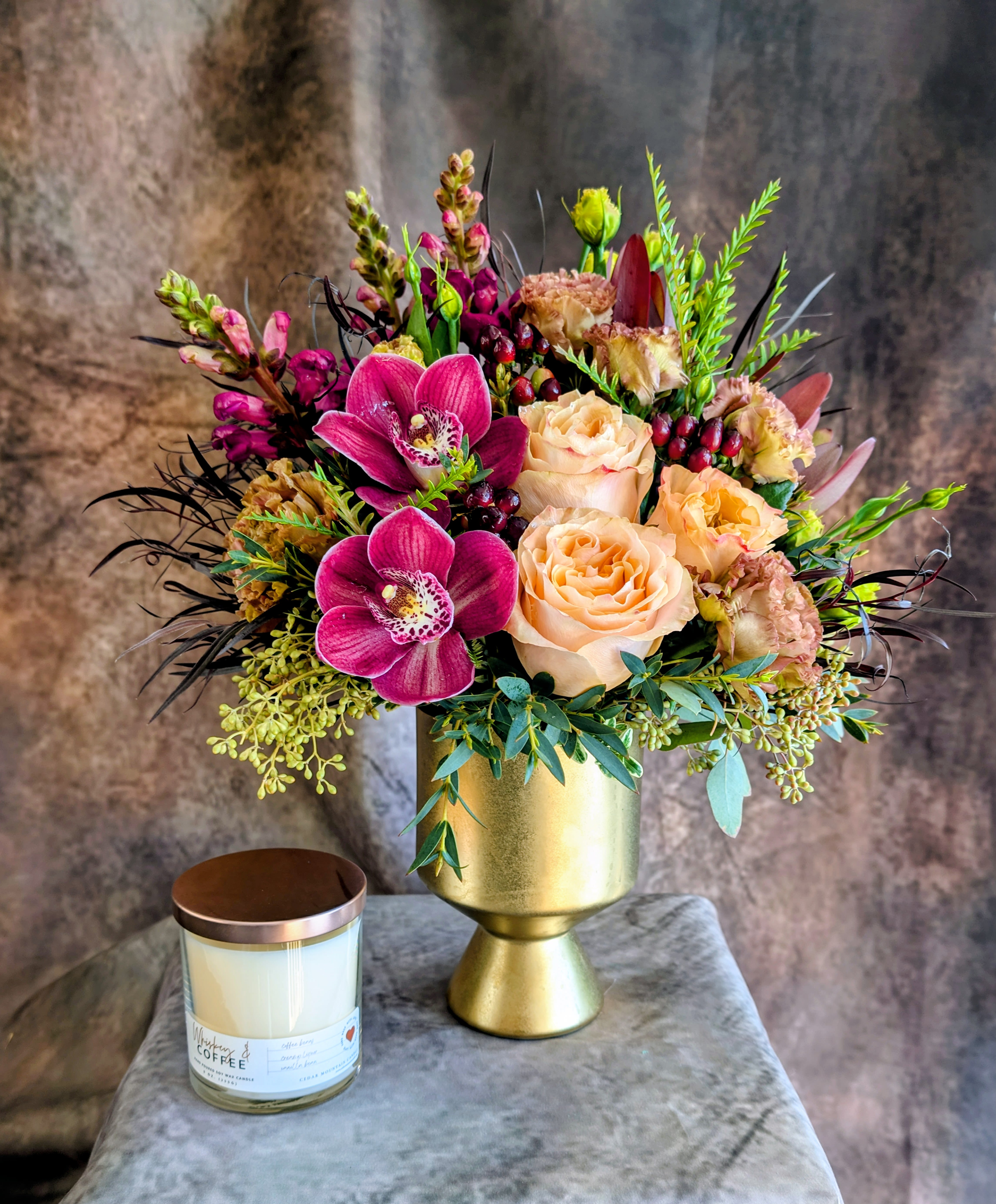 Brûlée- With A Cedar Mountain Whiskey And Coffee Candle - Introducing Brûlée, a warm and luxurious floral creation from Windmill Floral Expressions. This handcrafted arrangement blends glowing peach roses, velvety plum orchids, rich berry accents, and sculptural greenery—artfully gathered in a modern matte-gold pedestal vase. Inspired by the rich, caramelized tones of its namesake, Brûlée brings an elevated warmth and soft sophistication to any space. Paired with a decadent Cedar Mountain Whiskey and Coffee candle fragrance candle, this design becomes a full sensory experience—where the aroma of Irish coffee drifts through the room, perfectly complementing the arrangement’s rich jewel-tone palette. Thoughtful, modern, and indulgent, Brûlée makes an exceptional gift for the holidays, celebrations, or simply elevating your everyday home ambiance. Windmill Floral Expressions specializes in bespoke floral art, permanent botanicals, and mystical gifts—proudly delivering throughout Las Vegas, Henderson, Anthem, Inspirada, Seven Hills, and surrounding communities.   