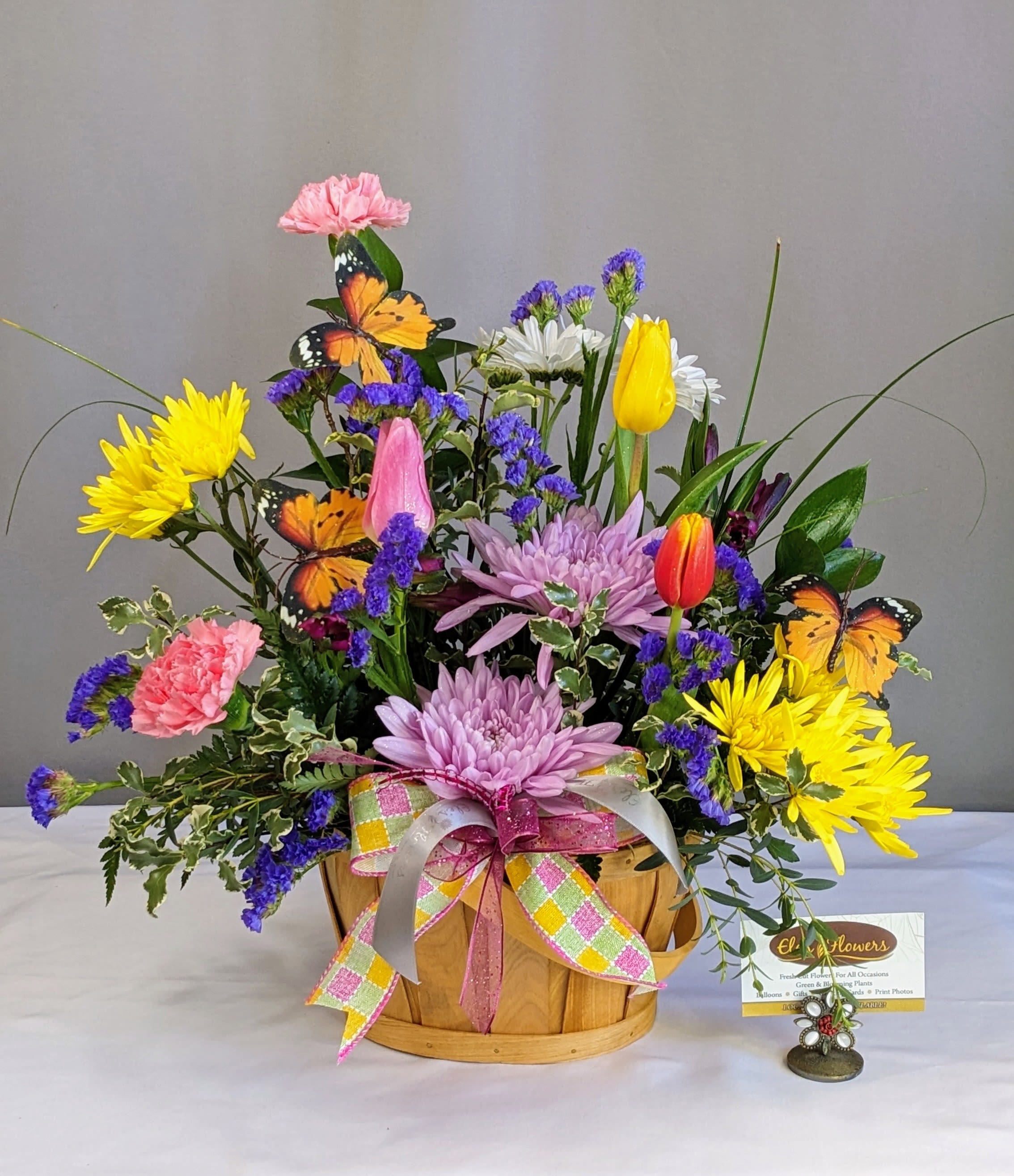 Jubilee Basket - Celebrate the joy of the season with our Jubilee Basket—a cheerful explosion of vibrant blooms that radiates warmth and happiness. This lively arrangement features a stunning mix of sunny yellow, pink and orange blooms, and pops of blue and purple accent flowers, all nestled in a charming wooden basket. Decorative butterflies bring a whimsical, garden-fresh feel, while a plaid pastel bow adds the perfect finishing touch. Perfect for any occasion, this arrangement is a burst of seasonal delight.   Blooms may vary from photo.    Approximate Dimensions: 18"H x 17"W