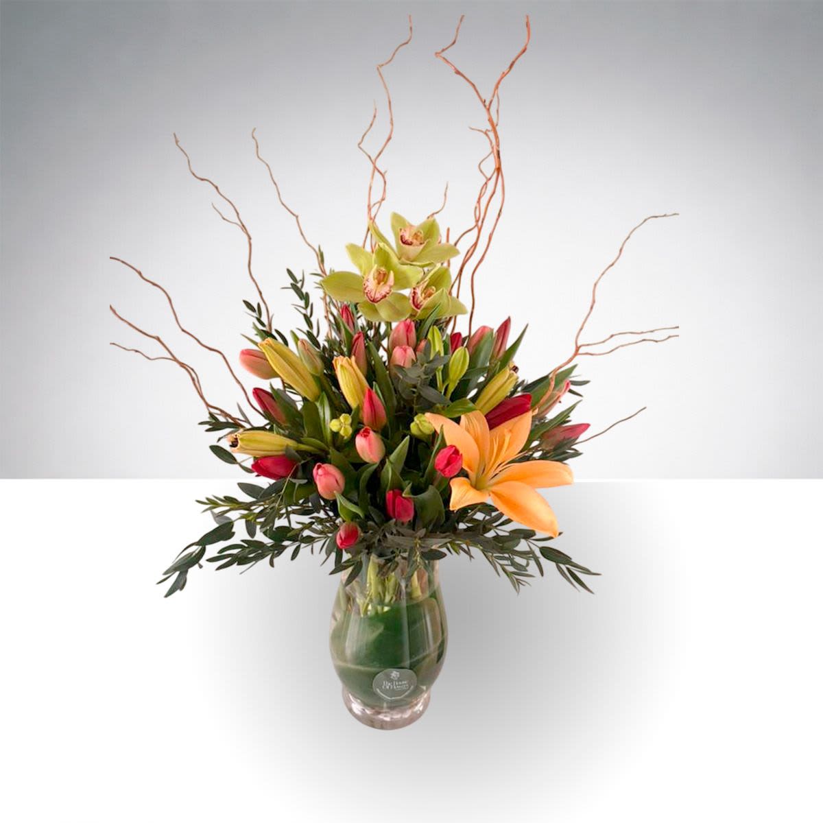 Sprng Graden Bloom by The House of Flowers - A vibrant and artistic floral arrangement featuring elegant orchids, fresh tulips, graceful lilies, and seasonal greenery. Designed with natural curly branches for height and movement, this arrangement creates a beautiful garden-style presentation in a clear glass vase.  The mix of warm colors and fresh blooms brings energy, beauty, and sophistication to any space, making it a wonderful gift or statement piece for special occasions.