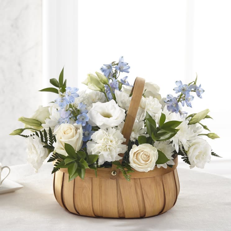 Sincerely Heartfelt Basket - A soft expression of comfort and grace. This charming wooden basket is overflowing with a serene collection of creamy white roses, ruffled carnations, and delicate lisianthus. Accented with airy blue delphinium and fresh garden greenery, it’s a gentle reminder of peace and love during life’s most meaningful moments.
