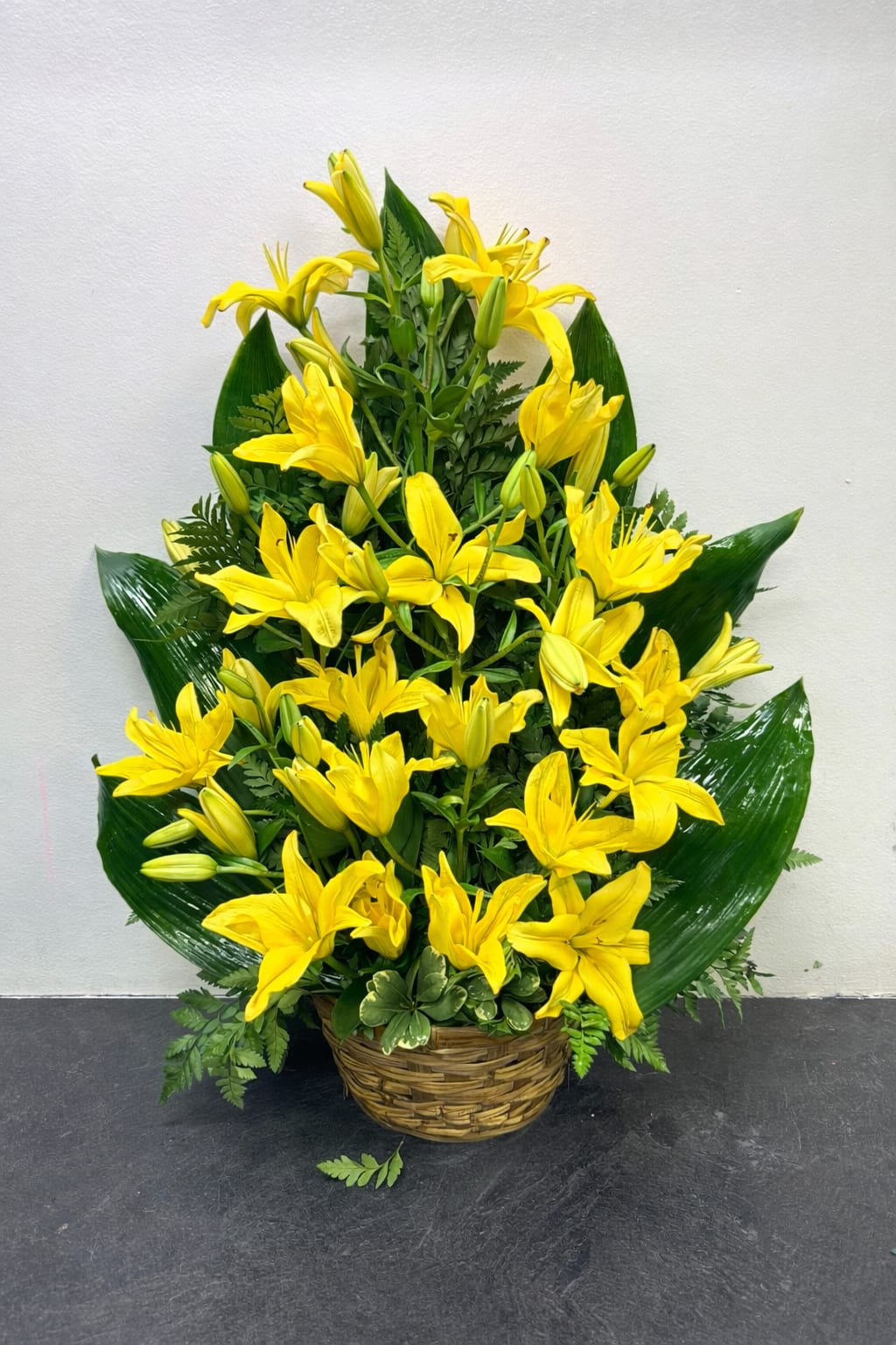 Sunrise smile  - A bold and uplifting arrangement featuring vibrant yellow lilies arranged in a full, cascading design. Each bloom opens gracefully, creating a sense of movement and brightness, while fresh green foliage adds depth and contrast. Set in a natural woven basket, this piece feels both elegant and grounded—perfect for celebrating life, sending encouragement, or bringing warmth into any space.  Radiant and expressive, it’s a statement arrangement that instantly captures attention and lifts the mood.
