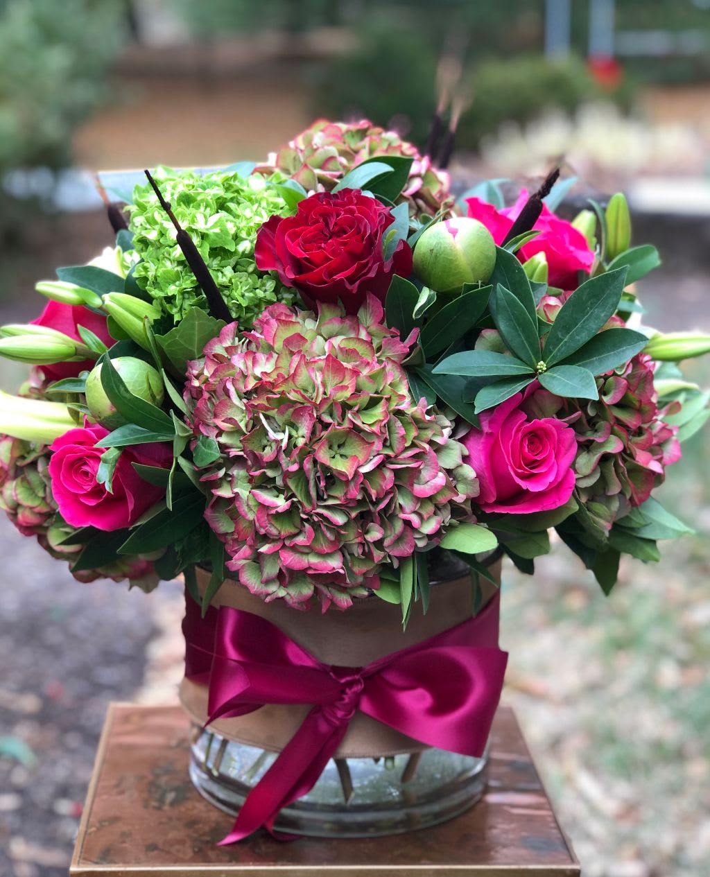 Heirloom Suede - Heirloom Suede | Wilmington, Delaware Florist  Discover rustic elegance with our Heirloom Suede floral arrangement, exclusively crafted by Petals Flowers and Fine Gifts — your trusted local florist in Wilmington, Delaware. This striking design showcases lush green and burgundy hydrangea, classic red roses, rich dark pink roses, graceful lilies, and unique cattails. Artfully arranged in a clear glass cylinder wrapped in natural burlap and finished with a sophisticated burgundy ribbon, Heirloom Suede brings warm, timeless beauty to any space.  Perfect for autumn gatherings, dinner parties, or simply elevating your home décor, this arrangement captures the charm of vintage-inspired florals with modern flair.  As a premier florist in Wilmington, Delaware, we specialize in custom, deluxe floral designs and guarantee every order with personal care. We proudly offer same-day, tax-free flower delivery throughout Wilmington, Hockessin, Newark, Glen Mills, Chadds Ford, Kennett Square, and nearby areas.  Why Choose Us?  * Locally owned boutique florist delivering to Wilmington and surrounding communities * Exceptional quality with an artistic eye for color and texture * Friendly, reliable service and a commitment to your satisfaction Order the Heirloom Suede arrangement today and let us bring a touch of refined beauty and natural texture to your special occasion or loved one’s doorstep in Wilmington, Delaware.