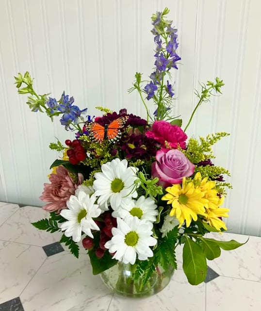 Garden Posy Party - A charming arrangement full of garden-inspired beauty. Garden Posy Party is designed in a classic “Rosie Posie” vase and features a delightful mix of daisies, carnations, chrysanthemums, and a soft lavender rose, finished with a delicate butterfly accent.  With a fresh color palette of purple, white, and yellow accented with touches of pink and red, this arrangement feels like a hand-picked bouquet straight from the garden. Cheerful, whimsical, and full of texture, it’s a perfect choice for celebrating life’s everyday moments.