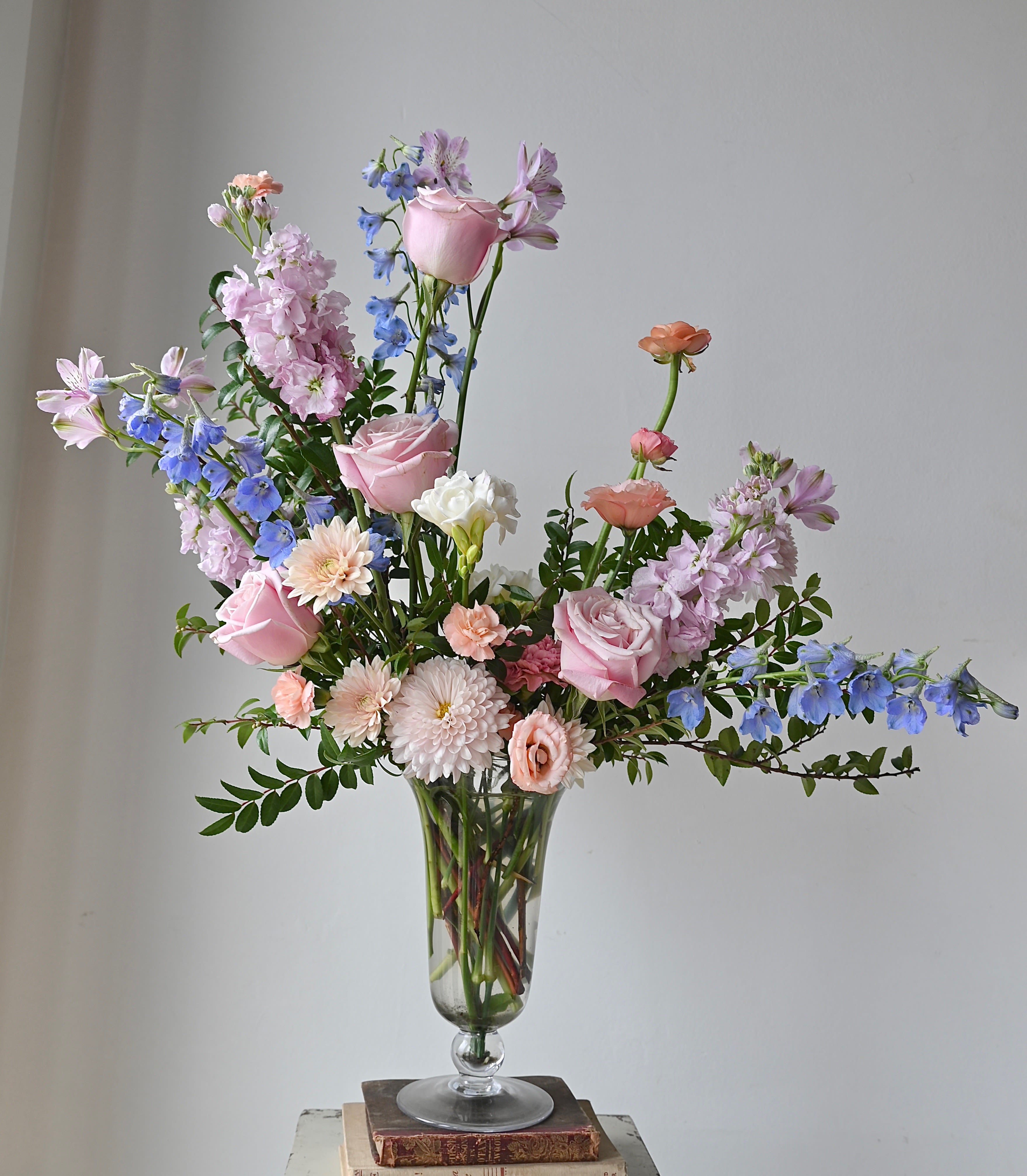 THE CAMBRIDGE - A true showstopper designed in an elegant footed glass vase.  A larger display of blooms and foliage; with a shape that  lends itself to lush, whimsical, garden-style design. Packaged in our signature white tissue and black logo ribbon.  Size Pictured: Basic  *Please note that our selection of flowers is always changing to ensure a fresh, seasonal mix of blooms. We cannot guarantee specific varieties and we will try to accommodate special requests.