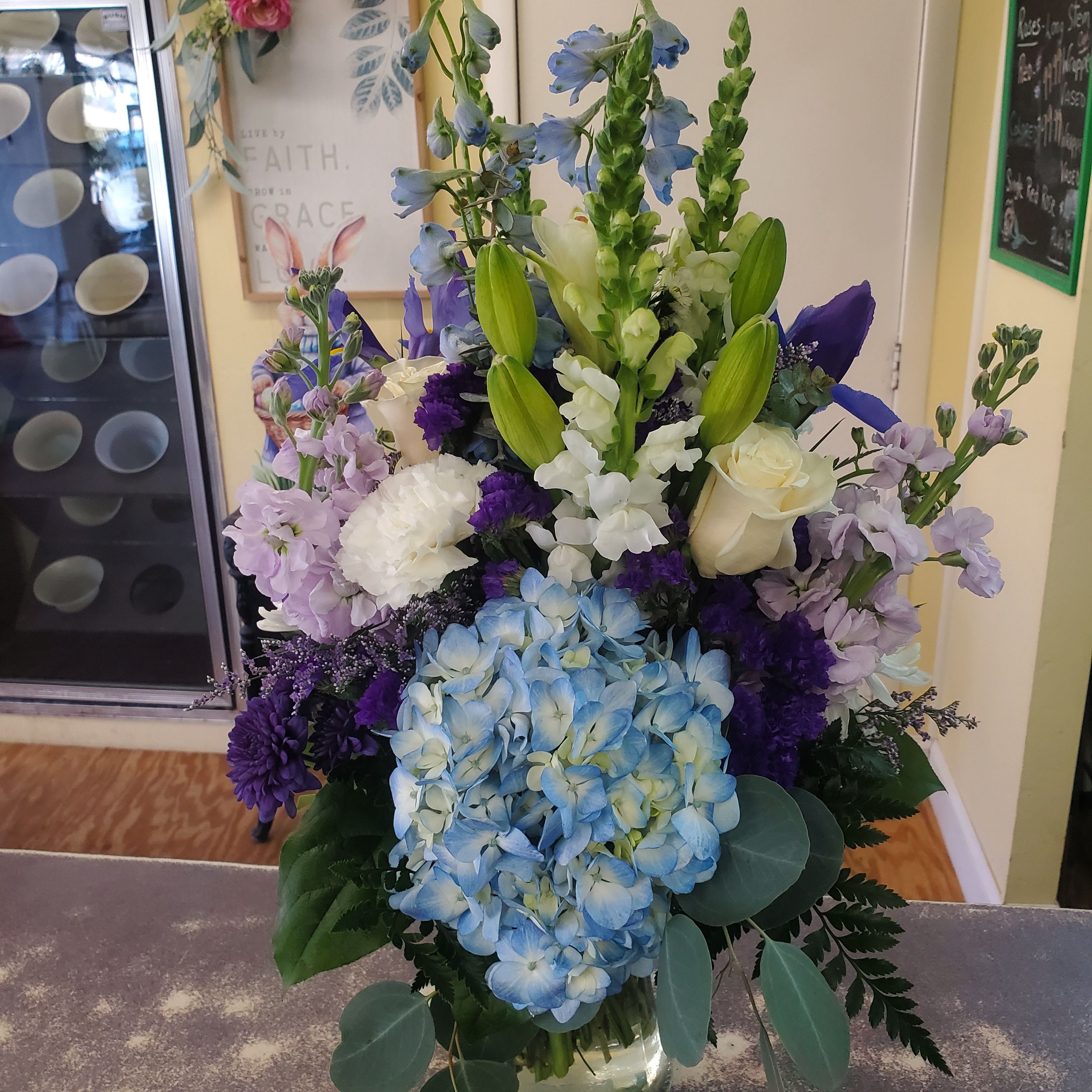  Blue Skies Bouquet  - Arrangement designed in a clear glass  container with assorted blues, purples and white flowers accented with assorted natural greens. 