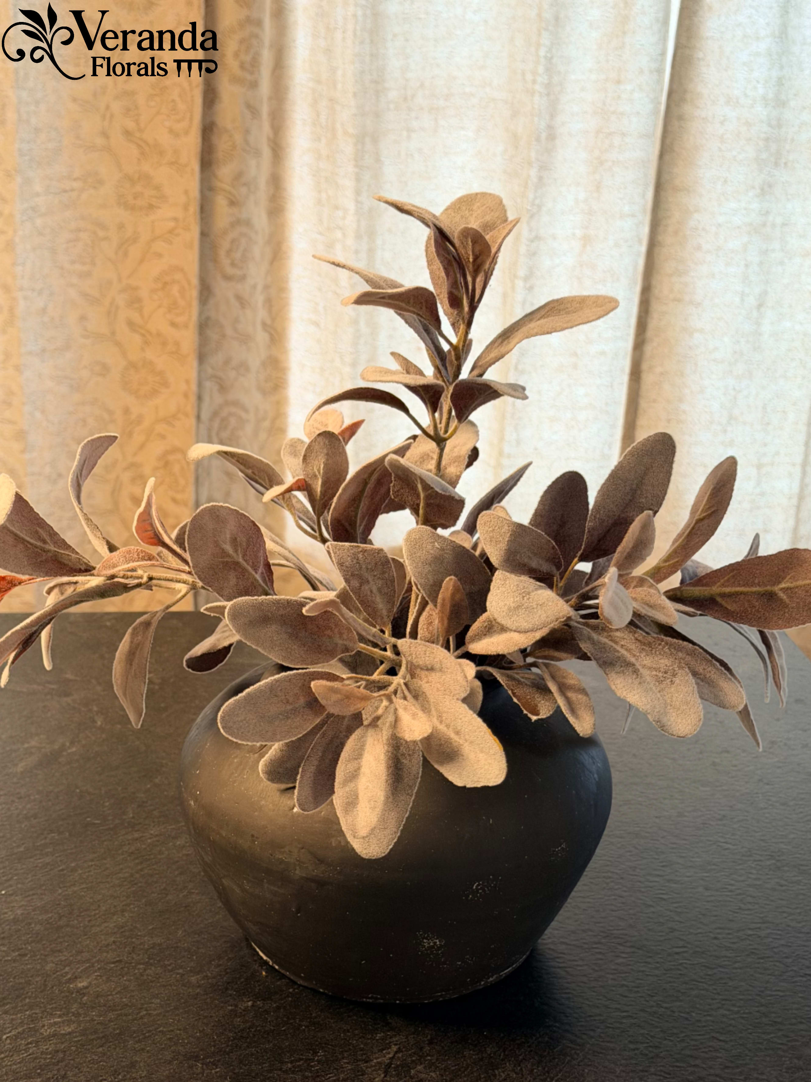 Muted brown faux foliage in a round black vase on a dark surface