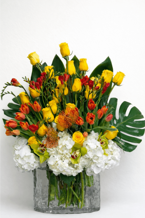 Tall arrangement of yellow roses, orange tulips, protea, orchids and white hydrangeas in a clear glass vase