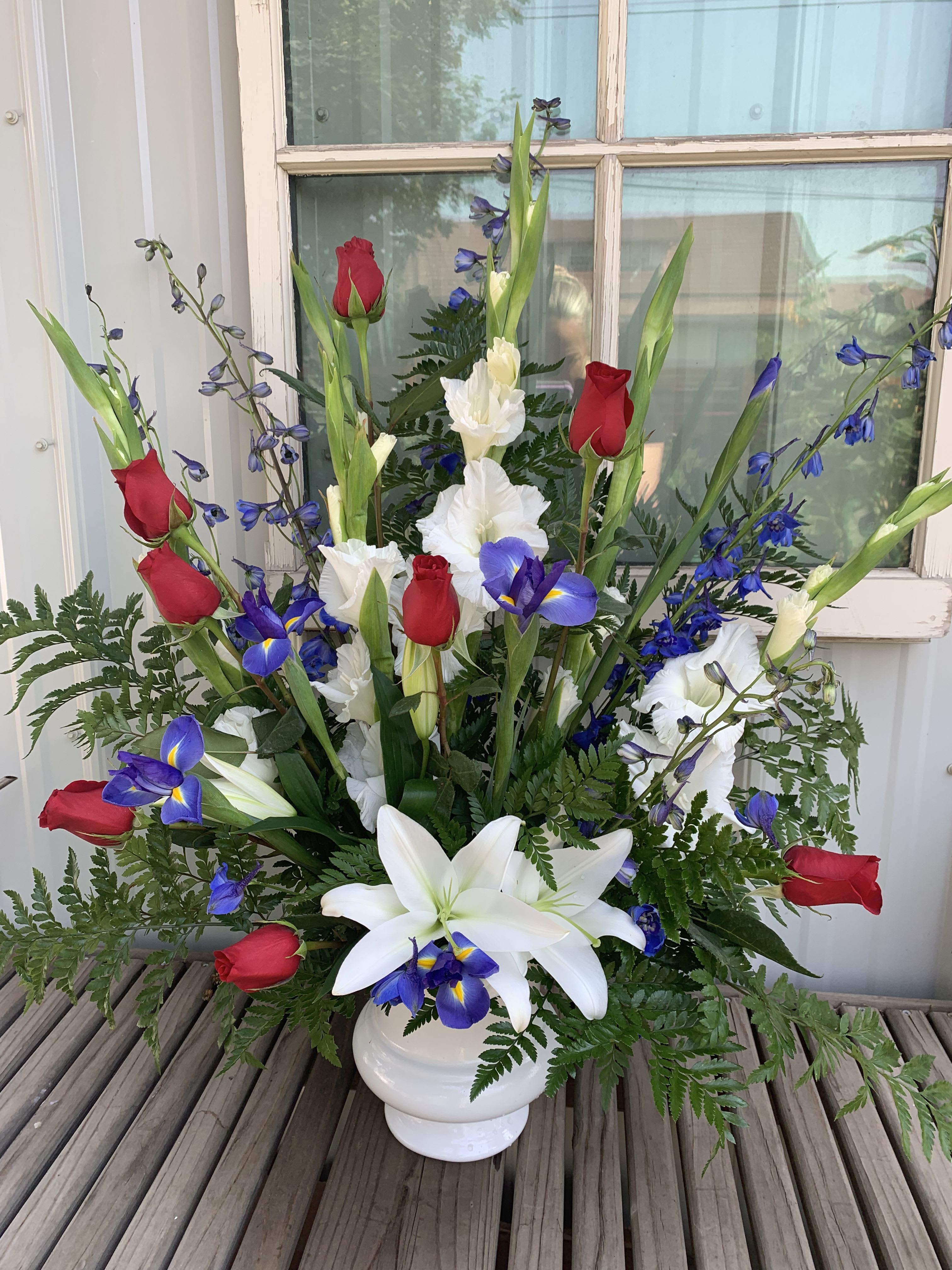 Cherished Farewell Arrangement - Cherished Farewell Arrangement is an elegant display of patriotic beauty to honor your loved one at their final farewell service. Red roses, white gladiolus, light blue delphinium, blue iris, white Oriental lilies, and lush greens are elegantly arranged in a white plastic urn to create a stunning way to say your last goodbye.