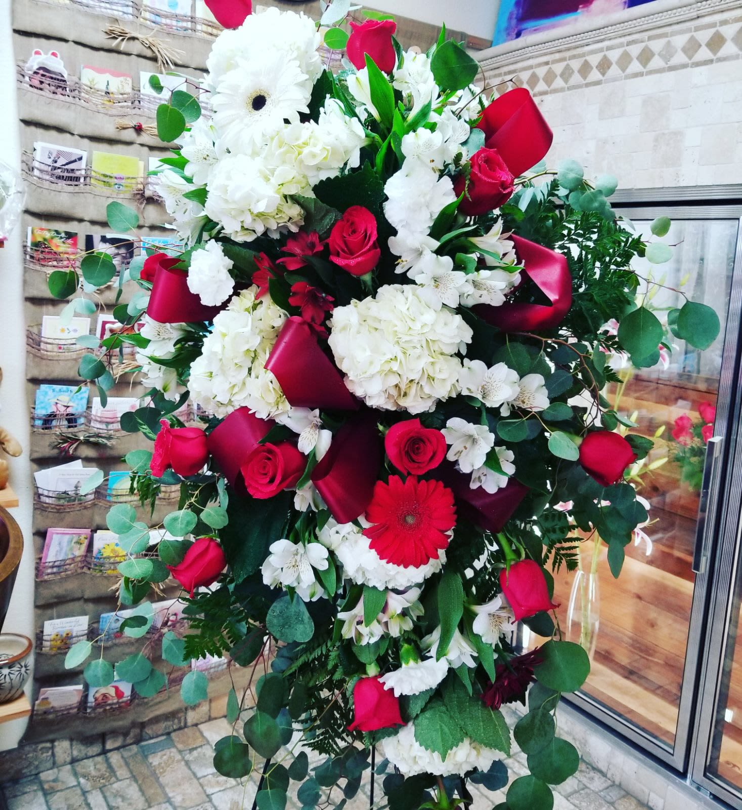 Red and white standing spray - A timeless expression of love, respect, and remembrance, this elegant standing spray features a classic blend of red and white blooms that convey both passion and peace. Beautifully arranged on a standing easel, the design showcases lush red roses and carnations symbolizing love and devotion, paired with pure white lilies, chrysanthemums, and roses representing reverence and serenity.  Accented with fresh greenery such as salal, leatherleaf fern, and eucalyptus, this graceful spray creates a stunning vertical display. A satin ribbon (available in matching red, white, or custom colors) adds a final touch of elegance.