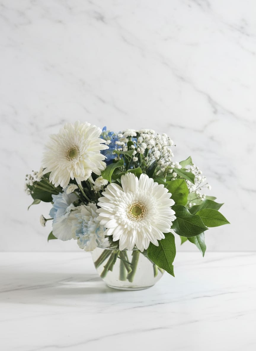 Misty Morning - A serene and elegant arrangement, the Misty Morning combines blue hydrangea and white gerbera daisies for warming combination, including gypsophila, whit roses, and green folliage in one transparent vase. Un arreglo sereno y elegante, la Mañana Brumosa combina hortensias azules y margaritas gerbera blancas para una combinación cálida, incluyendo gypsophila, rosas blancas y follaje verde en un solo jarrón transparente.