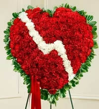 Broken Heart Carnation Tribute - A powerful and heartfelt standing spray featuring a solid heart of vibrant red carnations, accented with a white break to symbolize a broken heart. Lush fresh greens frame the design, while a coordinating red bow adds a polished finishing touch. This emotionally striking arrangement conveys love, remembrance, and deep sentiment in a bold, memorable presentation.