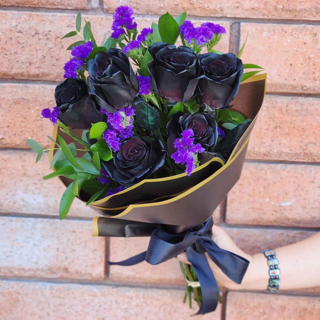 Black Rose Hand-Tied Bouquet - Black Rose Hand-Tied Bouquet – Premium Black Roses in San Diego  Make a bold, unforgettable statement with our Black Rose Hand-Tied Bouquet, featuring premium long-stem black roses accented with purple statice and lush seasonal greenery. Wrapped in black and gold designer paper with an elegant ribbon, this bouquet exudes mystery and sophistication.  Please note: Black roses are not naturally black; these roses are color-dyed to achieve their dramatic, striking hue.  Perfect for anniversaries, birthdays, gothic-inspired celebrations, or unique gifts, these rare, dramatic roses stand apart from traditional floral arrangements.  Pick-Up Only: Available for pick-up at our San Diego location.  Available Sizes:  • Standard: 6 Black Roses (color-dyed), Purple Statice Accents, Fresh Seasonal Greenery - Hand-wrapped  • Deluxe: 12 Black Roses (color-dyed), Purple Statice Accents, Fresh Seasonal Greenery - Hand-wrapped  • Premium: 24 Black Roses (color-dyed), Purple Statice Accents, Fresh Seasonal Greenery - Hand-wrapped  Care: Stems require a fresh cut before placing in water. Place in fresh water within a couple of hours of pick-up for best results.  Order your pick-up today and make a lasting impression with these stunning black roses!