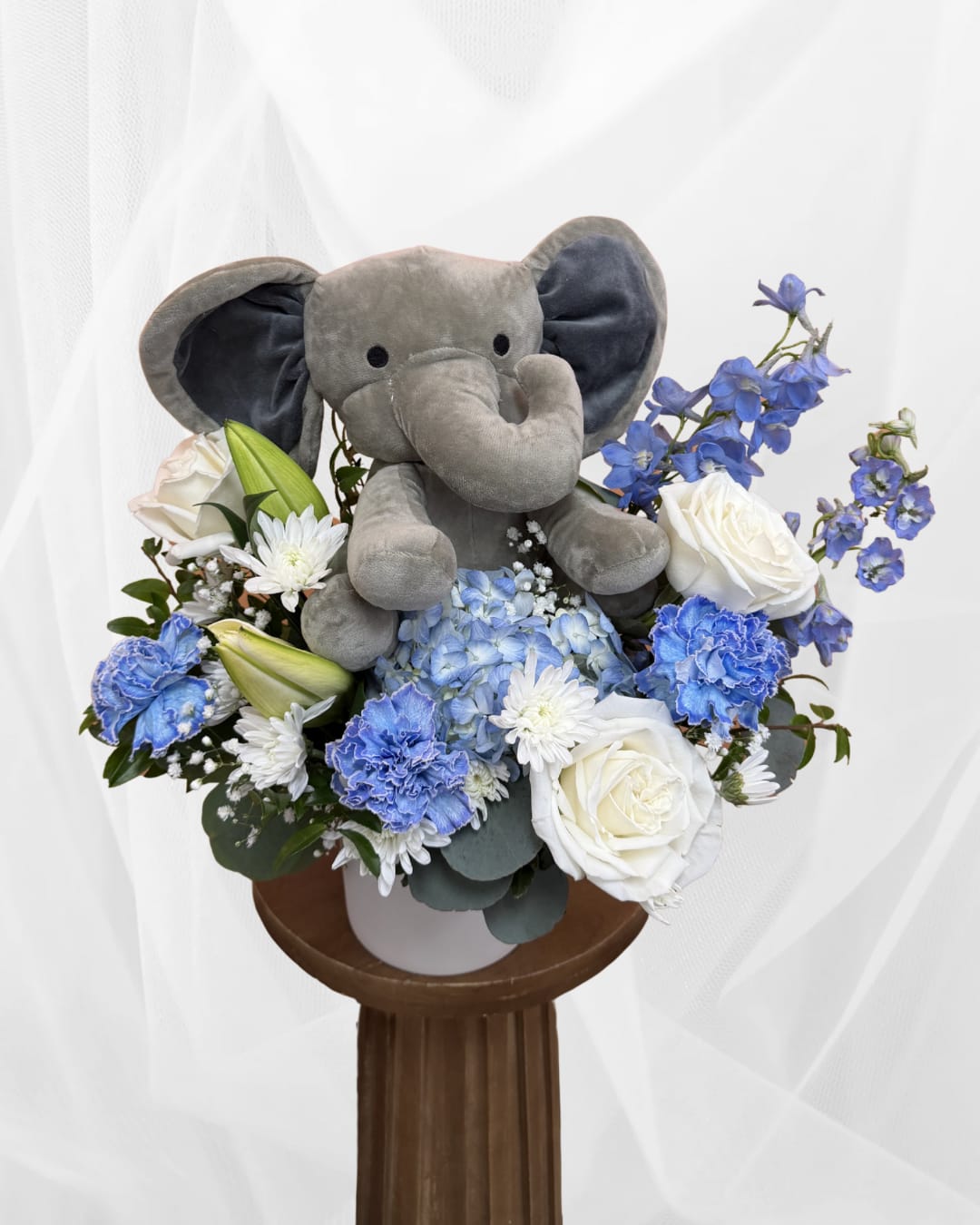 Baby Noah's Elephant Garden - Celebrate the arrival of a sweet baby boy with a charming floral arrangement designed to welcome new life with warmth and joy. This design features soft blue hydrangea, classic white roses, and delicate accent blooms arranged in a keepsake container alongside an adorable plush elephant. Aprox. dimensions 16x16