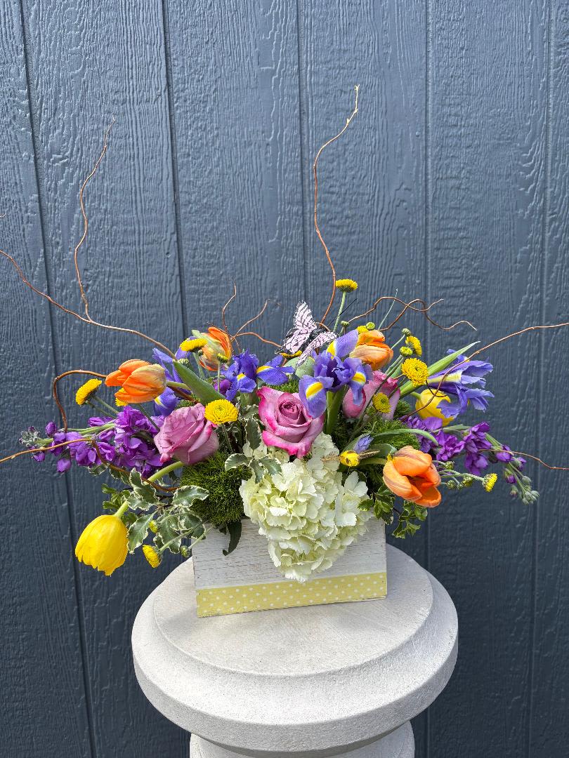 Bloom &amp; Flutter - Bloom &amp; Flutter  Light, airy, and full of movement, Bloom &amp; Flutter feels like a garden moment captured mid-breeze. Soft lavender roses and crisp white hydrangea are nestled among vibrant tulips, cheerful yellow accents, and delicate iris, creating a playful mix of color and texture. Whimsical curly branches dance above the blooms while a graceful butterfly adds the perfect finishing touch.  Designed in a charming keepsake container, this arrangement brings a fresh, joyful energy—perfect for celebrating spring, sending a smile, or brightening any space with a touch of nature’s magic.  Flower varieties and colors, Container colors may vary slightly based on seasonal availability.