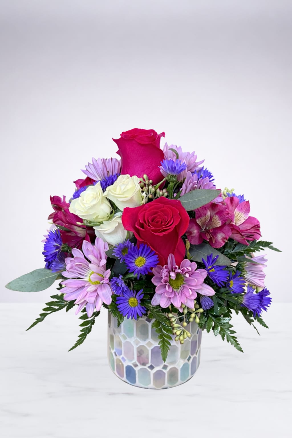 Blushing Garden Keepsake - A vibrant mix of hot pink and ivory roses paired with soft lavender daisies and pops of purple blooms, beautifully arranged in a mosaic vase. This compact design brings a cheerful, elegant touch to any space—perfect for celebrating life’s sweetest moments or simply brightening someone’s day.