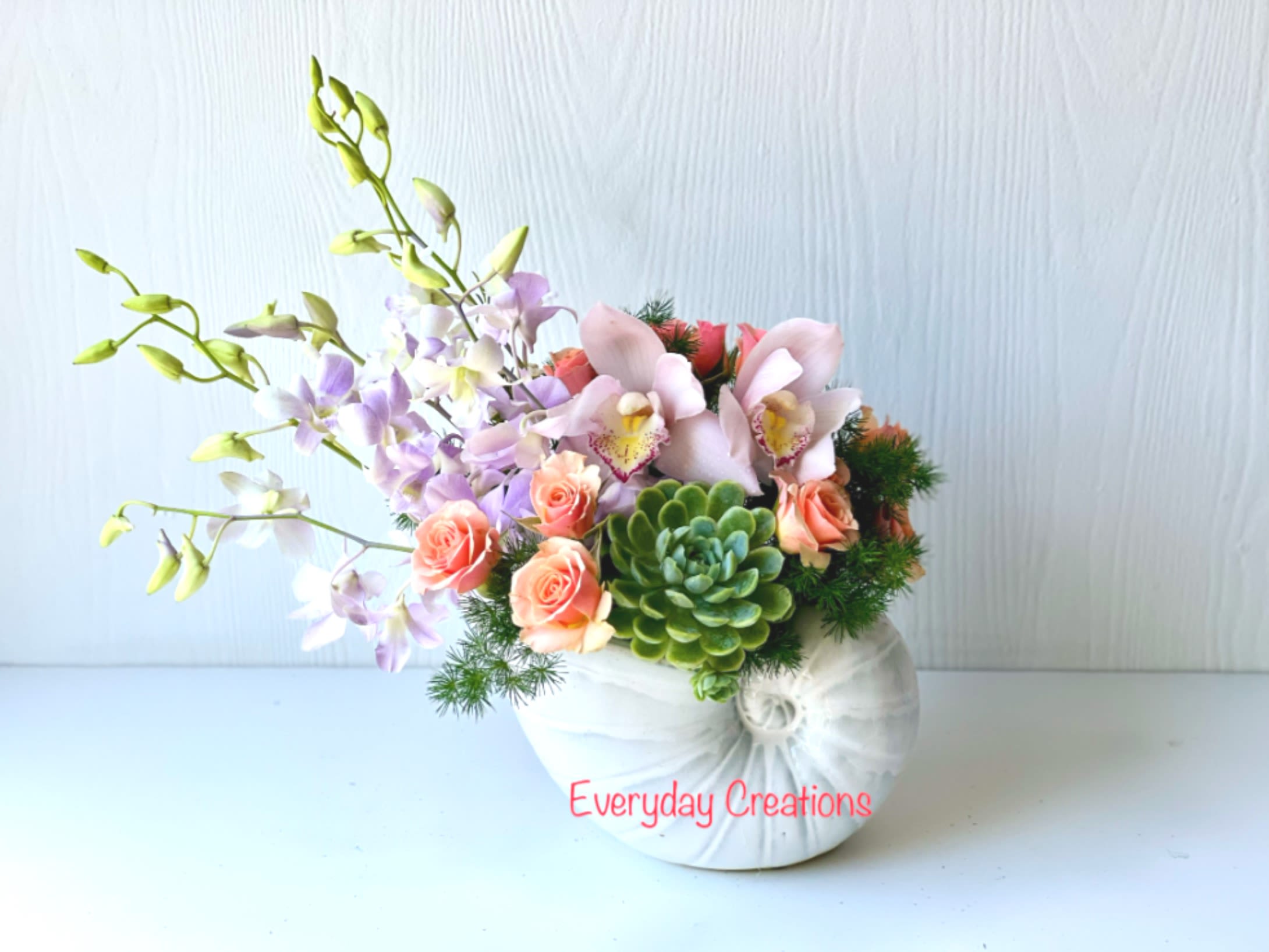 EC86 -  Coastal Charm  - Bring the tranquility and beauty of the beach into your space with this arrangement. This luxurious beachy floral arrangement features a stunning selection of premium flowers, including orchids, tulips, spray roses, succulents and a variety of greenery. All of the blooms are expertly arranged in a beautiful and unique shell vase to bring a sense of coastal charm.  Arrangement size: approx 10" wide, 13" high